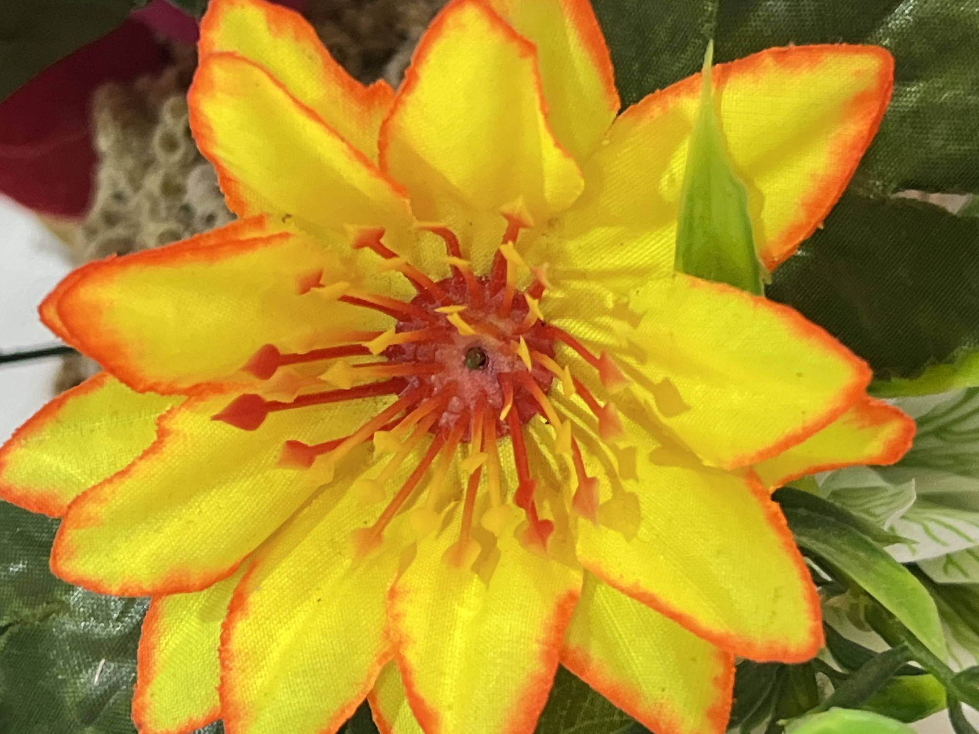 A close-up, head-on shot shows a bright yellow artificial flower with orange-tipped petals. The flower's center features multiple red stamens tipped with yellow. The background is blurred, with hints of green foliage and a textured, light brown surface. This image is not in Denpasar, Indonesia.