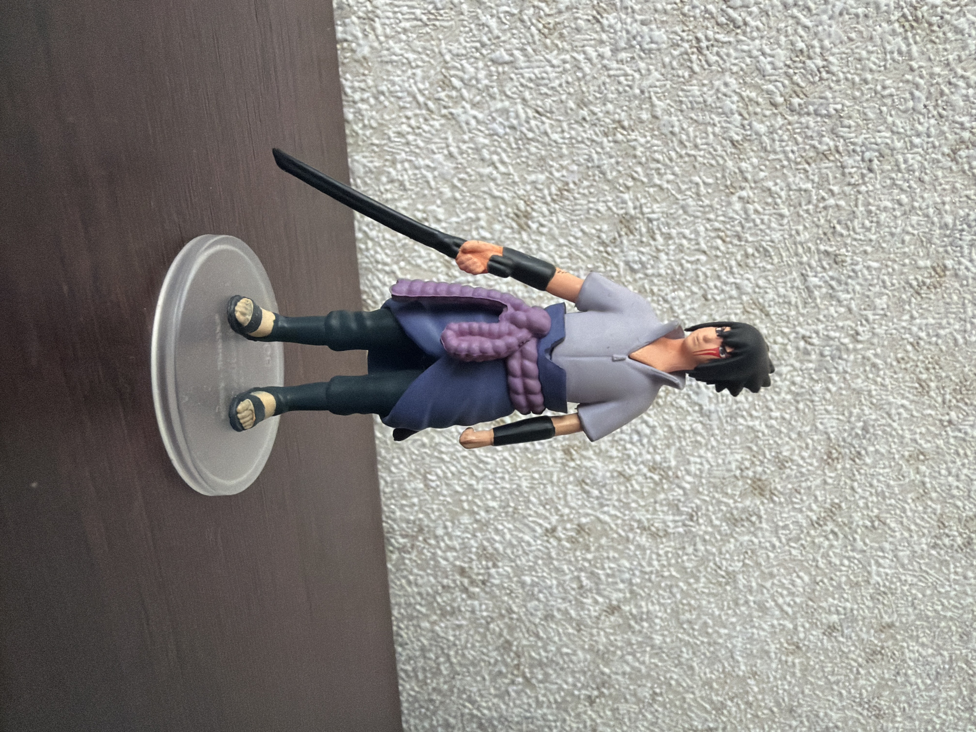 A full-body, eye-level, medium shot shows a Sasuke Uchiha action figure standing on a transparent circular base. The figure is positioned slightly off-center to the right. Sasuke is depicted wearing his typical outfit, consisting of a light purple, high-necked shirt, a dark blue loincloth tied with a purple sash, and dark pants. He has black hair, and his eyes are red with black sclera, and his face appears to have some red markings around the eyes. He is holding a black sword horizontally in his right hand. The figure stands on a dark brown wooden surface, with a textured off-white wall in the background. The lighting is even, and the focus is sharp on the figure.