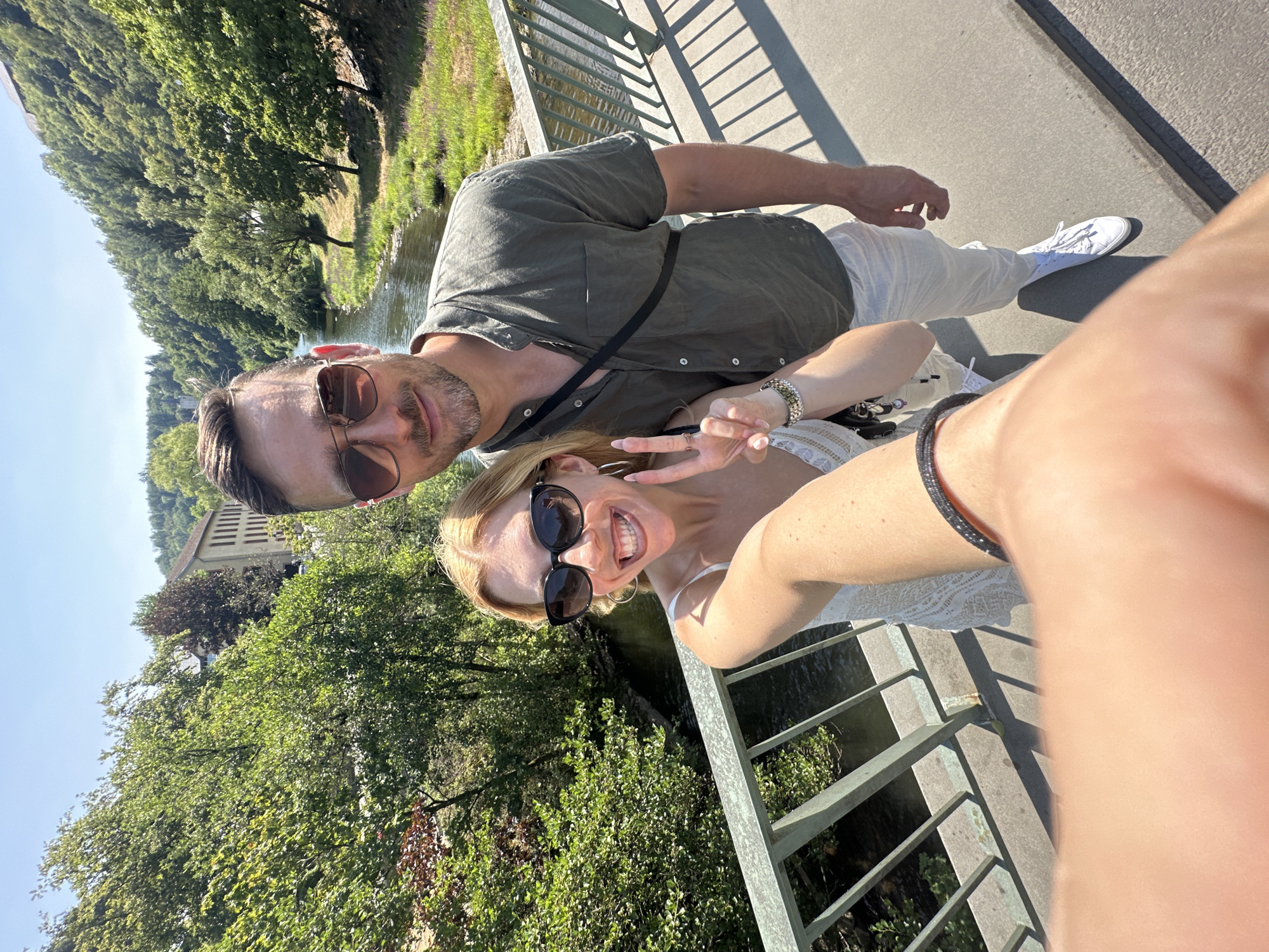 A man and a woman are taking a selfie on a bridge with a river and trees in the background, possibly in Vianden, Luxembourg. The woman, in the foreground and to the left, is wearing a white dress and sunglasses, and is making a peace sign with her right hand. She has blonde hair and is smiling. The man, to her right, is wearing a green shirt and sunglasses. He has brown hair and a beard, and is looking at the camera. The bridge has a metal railing. The river flows through a valley lined with green trees. A building with a red roof is visible in the distance. The sky is clear and blue.
