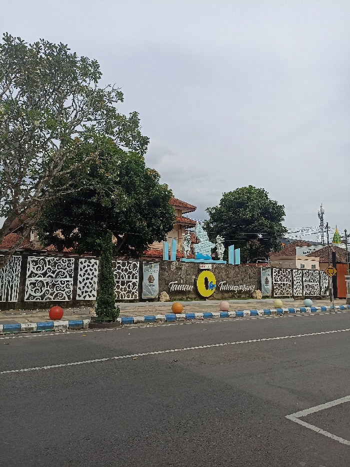 The image presents an outdoor scene depicting the entrance or monument of a public park in Tulungagung, Indonesia. The central feature is a low stone wall with the prominently displayed white text "Taman Tulungagung," indicating "Tulungagung Park." Above this, smaller blue text, likely "Selamat Datang di" (Welcome to), completes the message. A large yellow 'O' with a blue symbol inside is placed between the words "Taman" and "Tulungagung." On top of the stone wall, several light blue and white abstract sculptures are arranged.
In front of the wall, a decorative fence made of white metal panels with intricate swirling cutouts extends across the scene. Some dark plaques or banners are attached to these panels, but their text is unreadable. A slender, conical evergreen plant stands in front of the wall. Along the roadside curb, a series of colorful spherical bollards—red, orange, light pink, light blue, and yellow—are visible.
The foreground consists of an asphalt road with white lane markings and a distinctive blue and white painted curb. No people or vehicles are visible, suggesting a quiet moment. Large, leafy green trees frame the left side and background of the park area. Behind the wall and trees, several buildings with reddish-brown tiled roofs are partially visible. On the far right, a tall cell tower or antenna structure stands against the sky.
The scene is set during the daytime, but the sky is uniformly grey and overcast, indicating cloudy weather and a lack of direct sunlight. The absence of strong shadows further reinforces the cloudy conditions. The overall atmosphere is calm and uneventful.