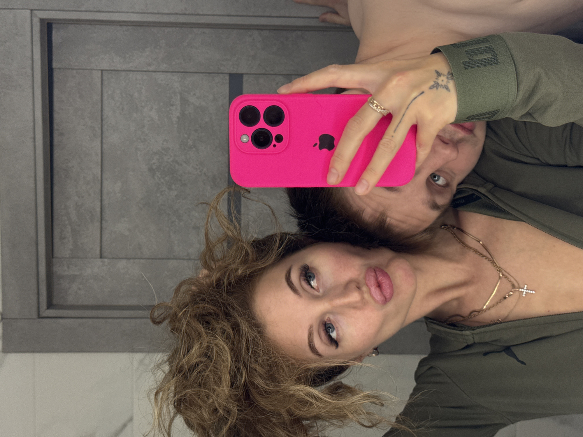 A selfie depicts a woman with curly brown hair and a man, both looking at the pink-cased iPhone held by the woman. The woman is pouting, and she is wearing gold necklaces. The man is behind her with a green, possibly Puma brand, zip-up jacket. The woman has a ring on her hand and a tattoo on her wrist. The background shows a grey patterned wall. The location is unknown and cannot be discerned to be Chaikovsky, Russia.
