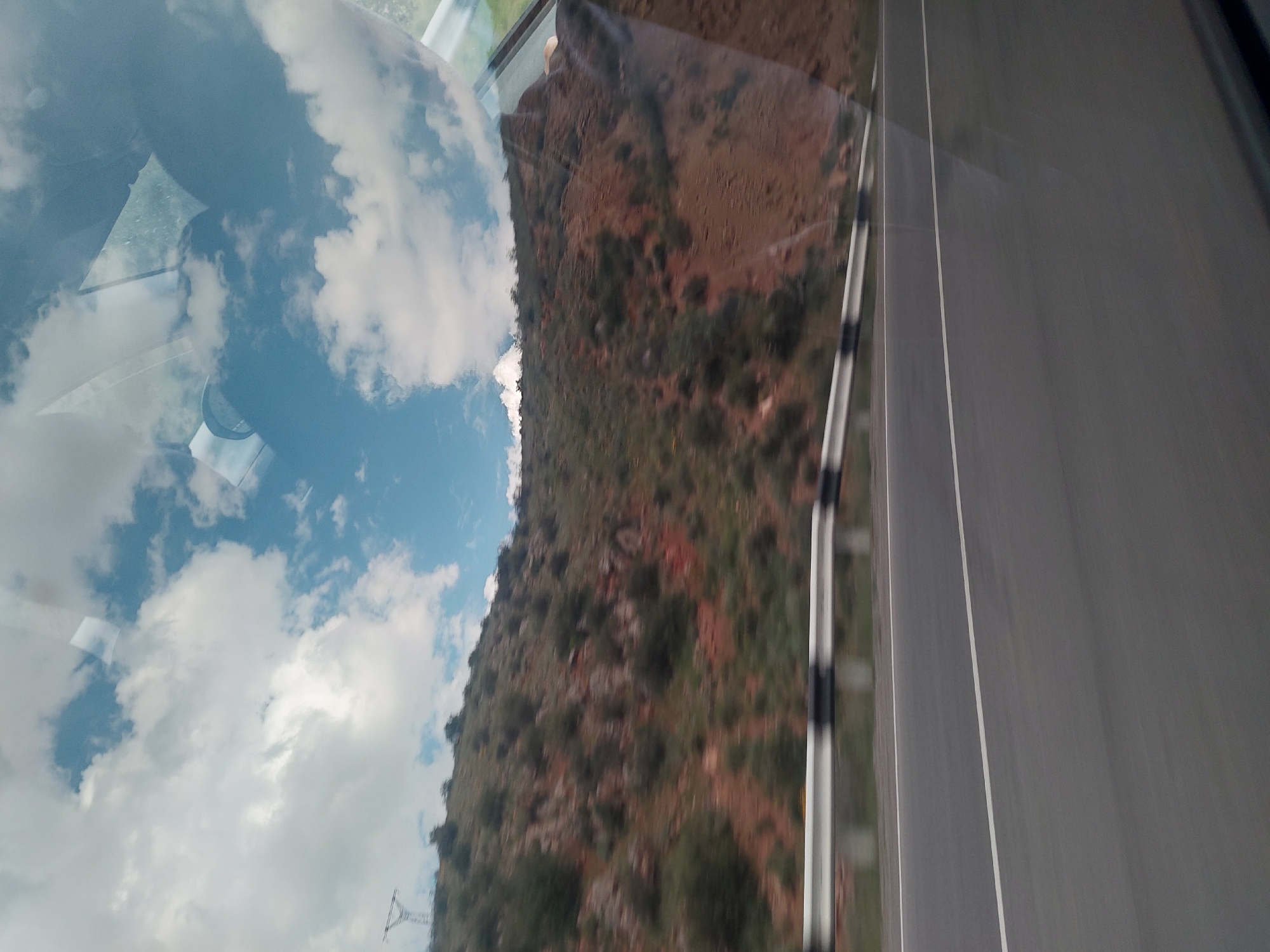 The image captures a scene viewed from inside a moving vehicle, looking out a side window. The photograph is oriented 90 degrees clockwise, causing the road to appear vertically on the right side of the frame.
On the far right, a dark asphalt road is visible, featuring two distinct white painted lines running parallel along its length. To the left of the road, a black and white striped guardrail separates it from a rugged mountainside. This mountainside, which dominates the central and left-center portions of the image, is characterized by reddish-brown soil and rocks, interspersed with sparse green and brown vegetation, primarily bushes and small trees.
The left side of the image displays a bright blue sky filled with large, white, fluffy clouds. This sky and cloud formation are also strongly reflected on the vehicle's window, particularly towards the far left, where they overlap with faint details of the vehicle's interior, possibly including part of a seat or a passenger's arm. A faint outline of a metallic lattice structure, likely a power line tower, can be seen against the sky in the lower-left quadrant.
The scene depicts a journey through a mountainous or hilly region during daylight hours under a partly cloudy sky. No discernible text is visible in the image. The location is Tigranashen, Armenia.