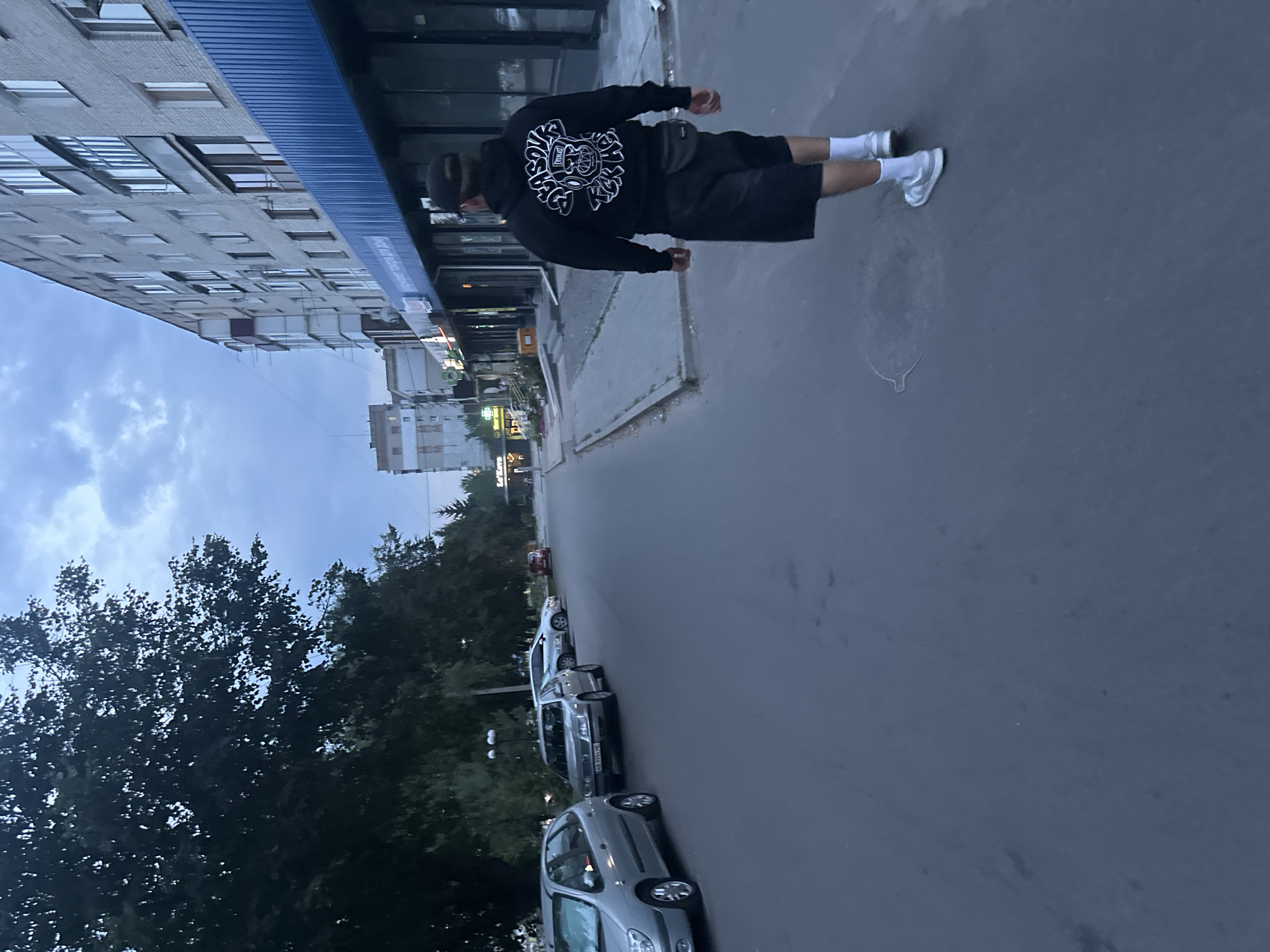 An eye-level, medium shot captures a person walking away from the camera down a paved street in Khmilnyk, Ukraine. The person, wearing a black hooded sweatshirt with a white graphic on the back, black shorts, white socks, and white and gray sneakers, carries a black fanny pack around their waist. The street is lined with parked cars on the left, including a silver Renault Scenic and a silver Volkswagen Passat. On the right, a multi-story building with a blue awning runs along the sidewalk. The sky above is overcast with a light blue hue. Trees with dark foliage are visible on the left side of the street. The scene appears to be set in the late afternoon or early evening.