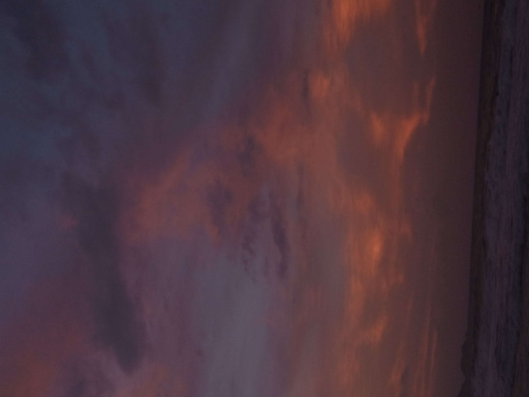 The image depicts a dusk or dawn scene at Playa Parguito, Venezuela. The sky is filled with clouds tinted in shades of purple and orange by the sunlight. Along the right edge is the coast with the water washing onto the shore.