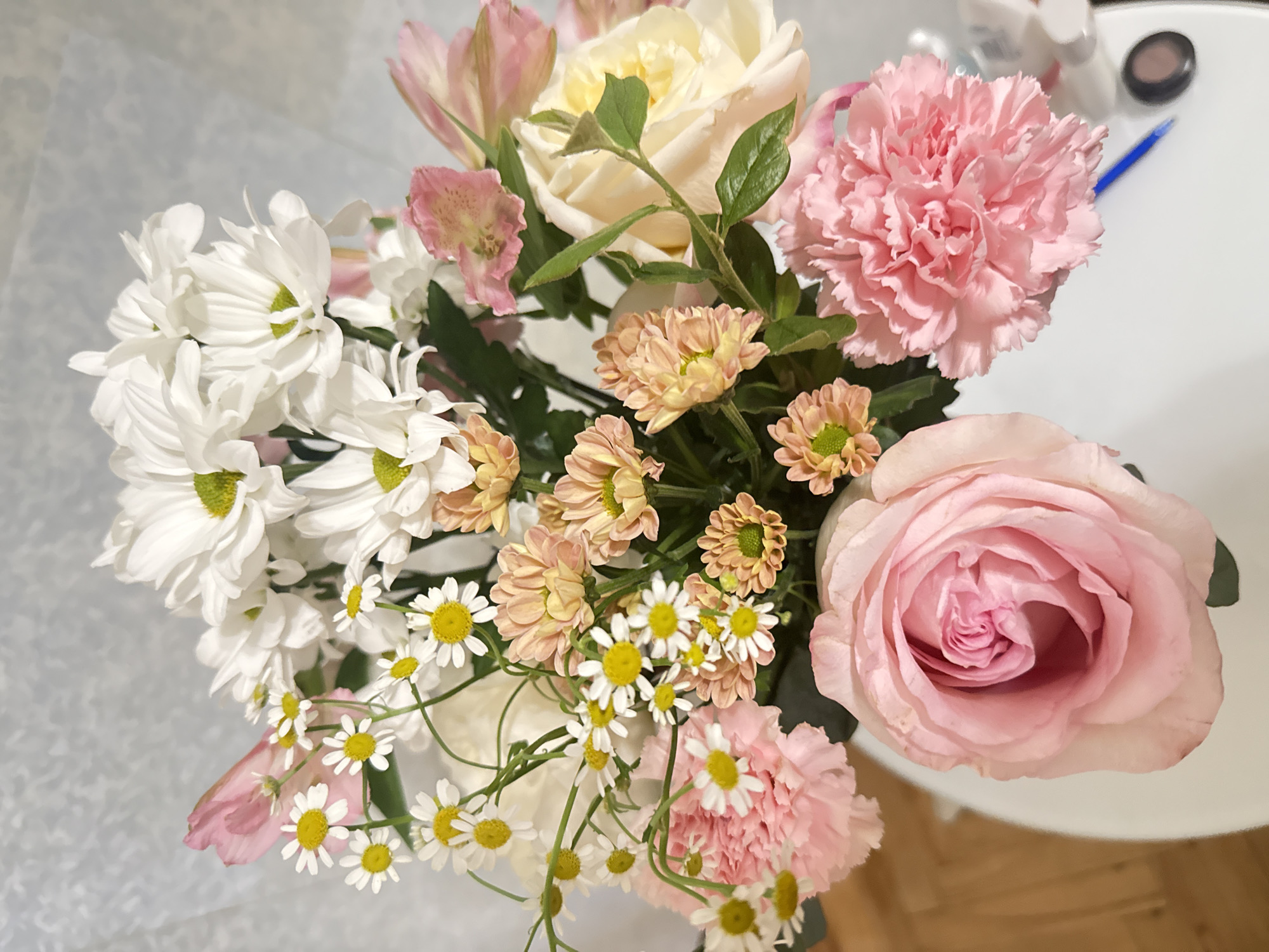 A close-up, overhead shot shows a bouquet of mixed flowers arranged in a white vase, resting on a white surface, likely a table. The bouquet features a variety of blooms including large, soft pink roses, ruffled pink carnations, white chrysanthemums with green centers, delicate white daisies with yellow centers, and clusters of small peachy-pink flowers. Some light pink alstroemeria petals are also visible. Green leaves and stems are interspersed throughout the arrangement. The background is a soft, textured white fabric. The scene appears to be indoors. There are no people or animals visible. The location in Saint Petersburg, Russia is not discernible from the image.