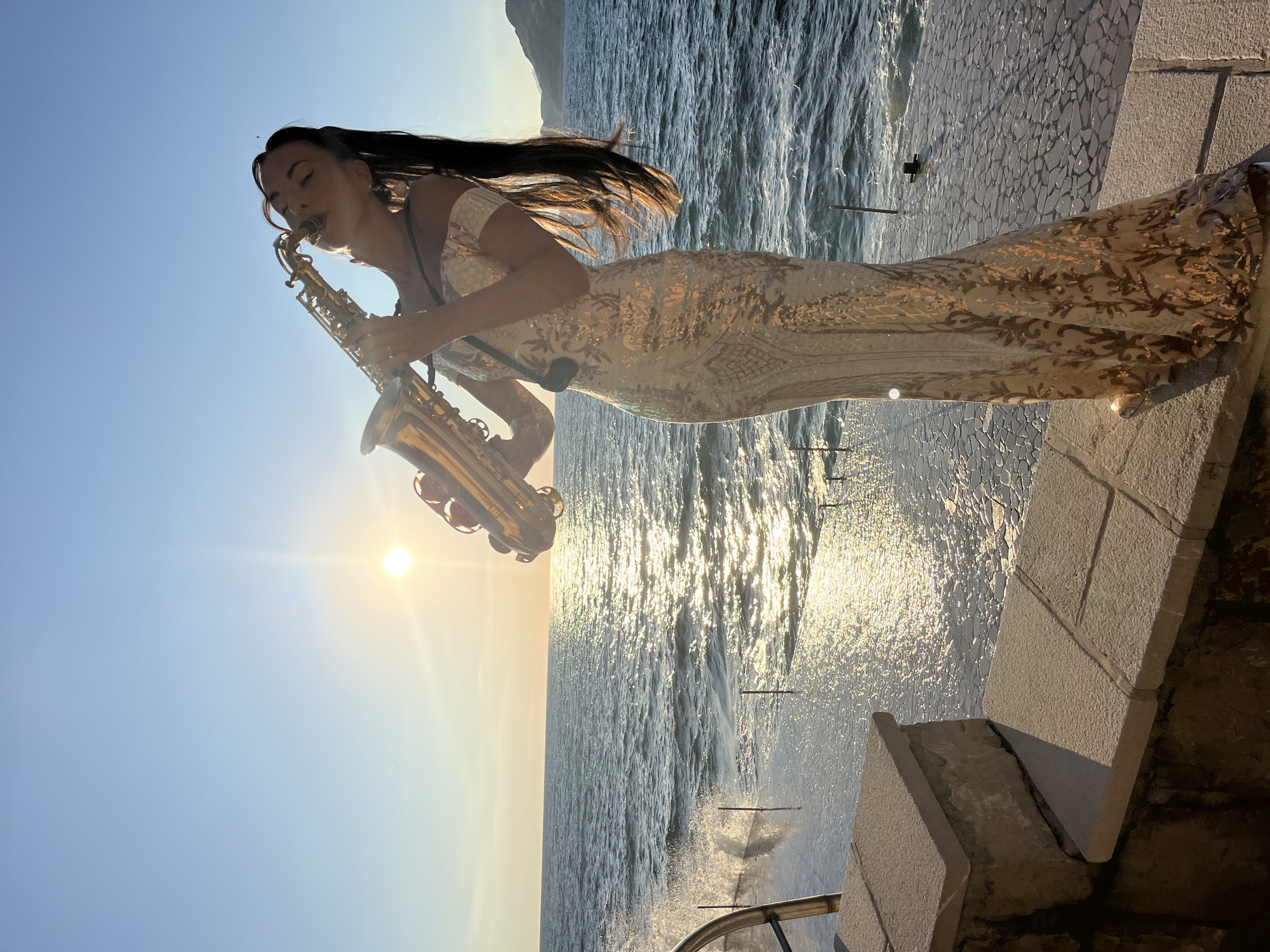 A woman plays a saxophone at sunset on a stone ledge overlooking the Adriatic Sea. The woman is wearing a long, off-the-shoulder, sequined gown. The sun is low in the sky, casting a golden glow on the water and the woman. Waves crash against the stone ledge. In the distance, mountains are visible. The location appears to be in or near Pečurice, Montenegro.
