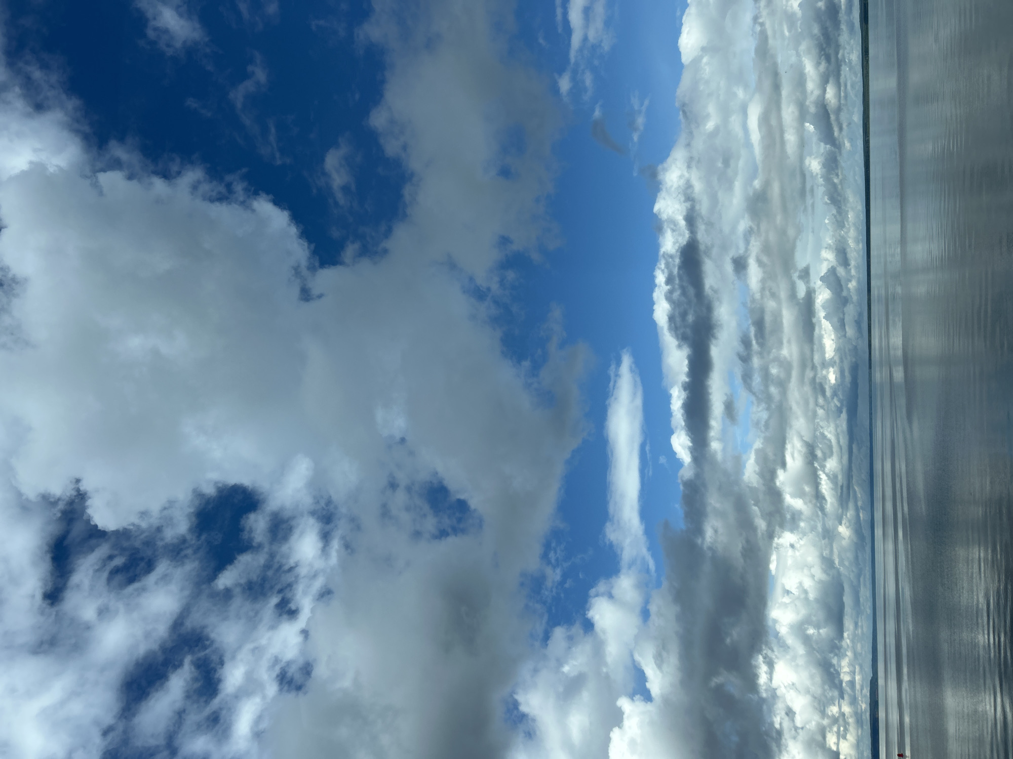 The image depicts a vast expanse of calm water under a dramatic sky filled with dynamic cloud formations. The water occupies the lower half of the frame, reflecting the muted tones of the clouds and the deep blue of the sky. The horizon line is visible in the distance, where the water meets a hazy, dark landmass. The upper half of the image is dominated by a complex interplay of light and shadow in the clouds. Bright white cumulus clouds are interspersed with darker, more ominous gray formations, creating a sense of depth and movement in the sky. Patches of clear blue sky are visible between the clouds, especially towards the top and middle of the frame. A small, red buoy is barely visible on the left side of the water, near the horizon. The scene evokes a feeling of open space and natural beauty. This image likely depicts the Kama River in the city of Chaikovsky, Russia, a region known for its expansive waterways and dramatic skies.