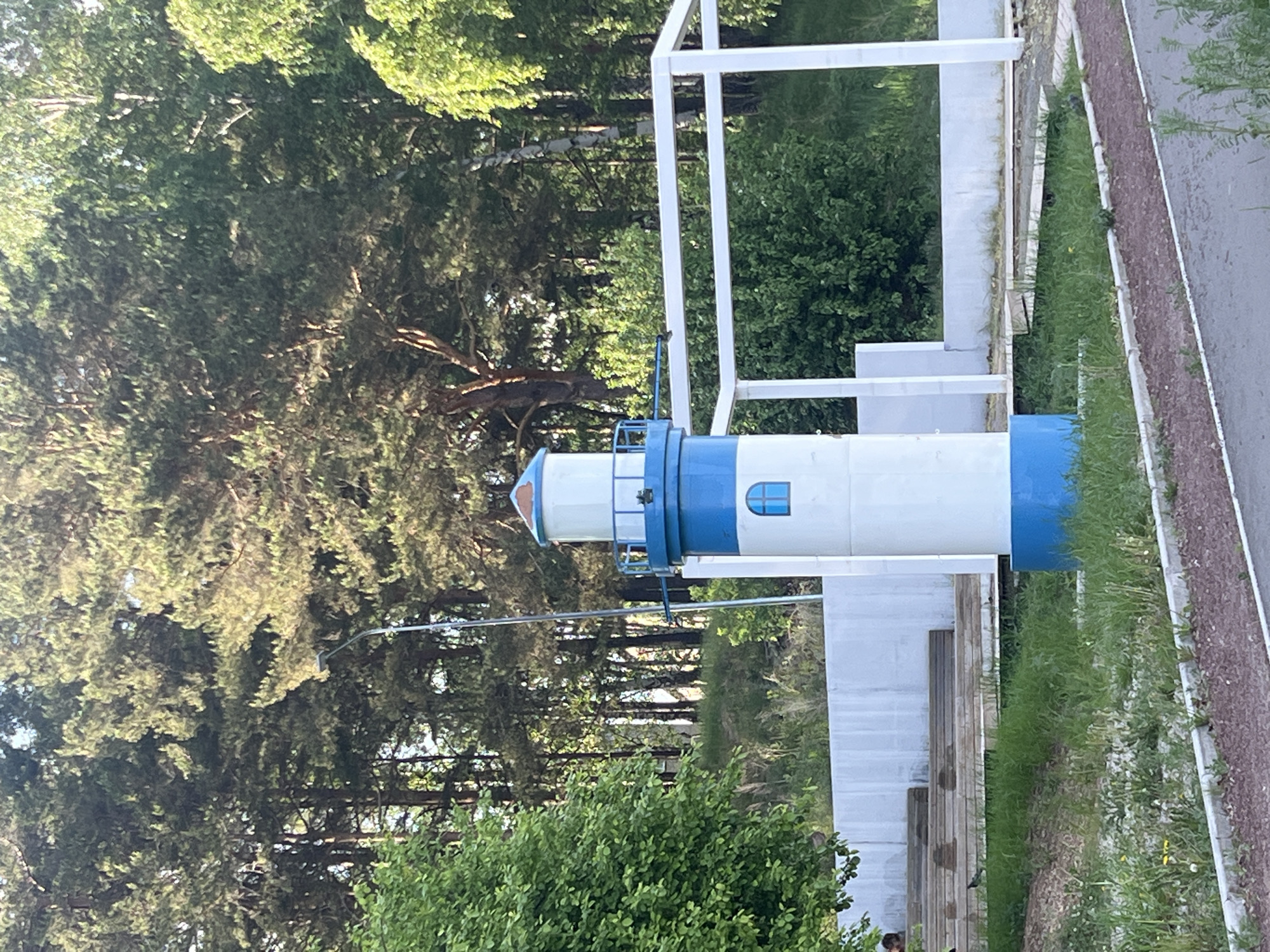 The image shows a white and blue decorative lighthouse structure with a viewing platform. It is located in a grassy area near a paved walkway in a park or recreation area. The backdrop is a forest with dense trees. The lighthouse is located in Chaikovsky, Russia. A metal utility pole is running overhead. There is some shrubbery to the front of the lighthouse.