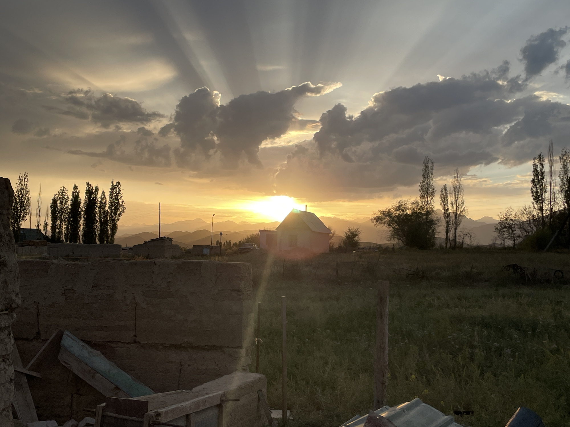 The image depicts a rural landscape at sunset in Kara-Oi, Kyrgyzstan. The sky is dramatic with crepuscular rays emanating from the setting sun, which is partially obscured by dark, billowy clouds. The sun is positioned low on the horizon, casting a warm, golden glow across the scene. In the middle ground, a small, light-colored house with a peaked roof is visible, silhouetted against the bright sky. A faint lens flare is present, suggesting the sun's intensity.
The foreground is dominated by a rustic, unfinished wall constructed of earth and stone, with some wooden planks and debris lying nearby. To the left of this wall, a pile of building materials, including what appears to be a metal sheet, is present. The middle ground features a grassy field with some sparse vegetation and a few wooden posts. Tall, slender trees are scattered throughout the landscape, particularly on the left side and in the background. A car is parked near the house.
In the far background, a range of layered mountains is visible, fading into a hazy orange sky. The overall impression is one of a serene, remote, and perhaps somewhat impoverished rural setting experiencing the beauty of a sunset. There are no people visible in the image.
