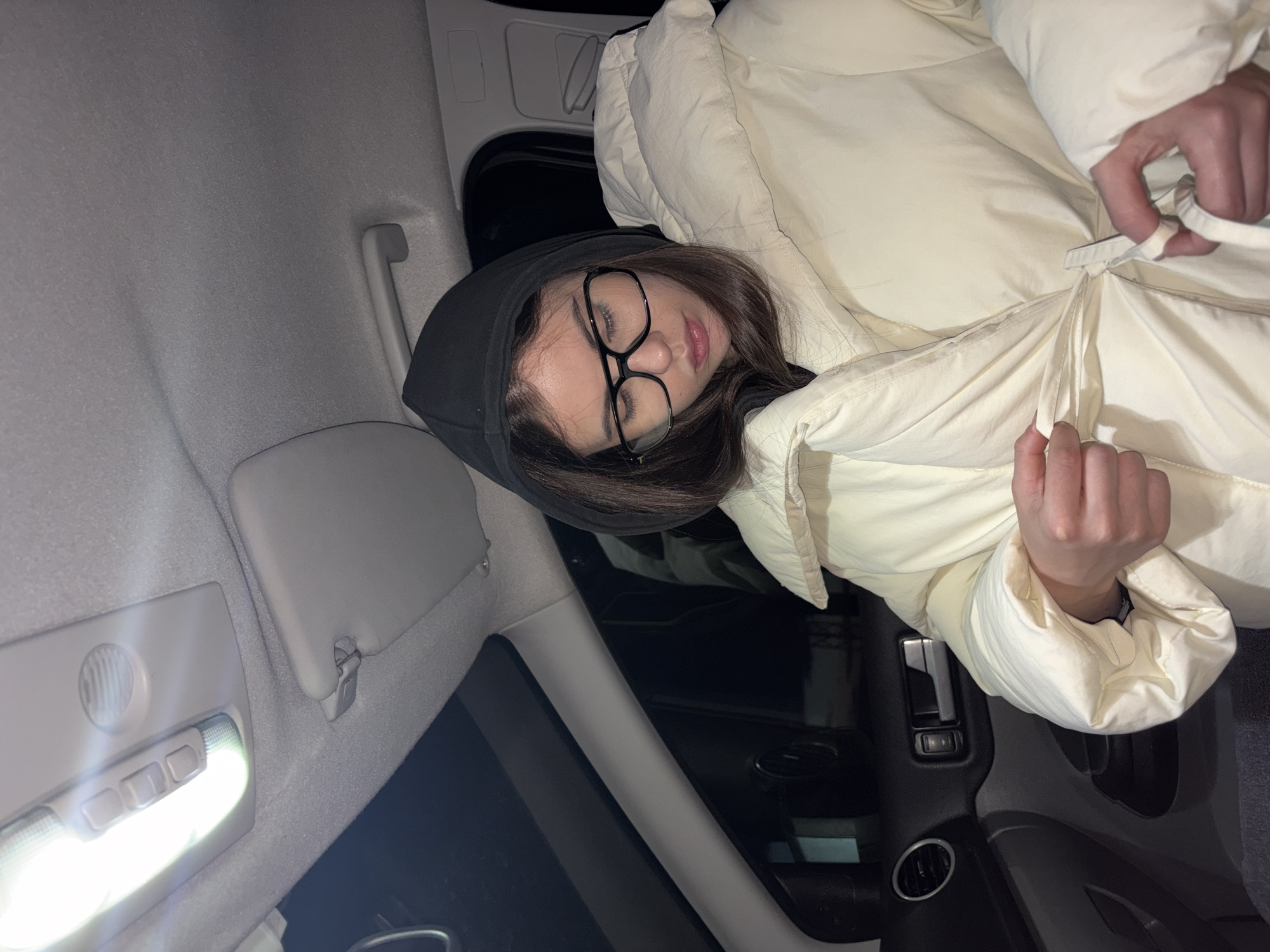 A woman with brown hair wearing glasses and a black hoodie is lying in the back seat of a car. She is wearing a cream-colored puffer jacket and appears to be sleeping. The car interior is visible, including the ceiling light, sun visor, and door frame. The location is unknown.