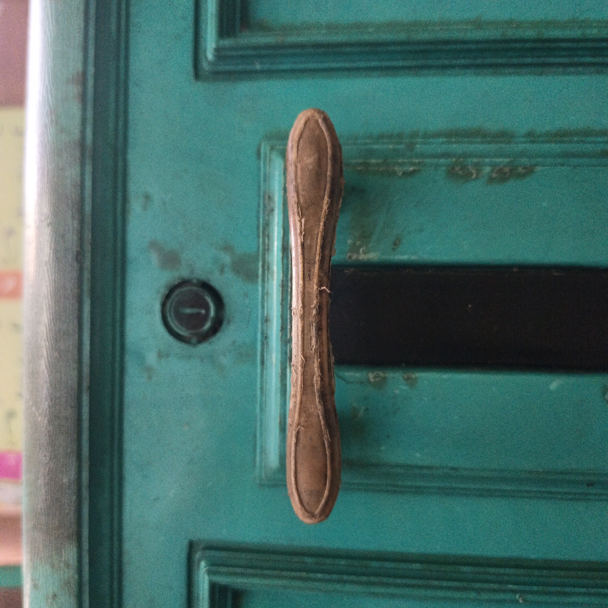 A close-up, eye-level shot shows a weathered, teal-painted door with a protruding, elongated, brown wooden handle. The handle is vertical and has a slightly worn, textured surface. To the left of the handle, a circular keyhole is visible, and to the right, a horizontal black band runs across the door. The door appears to be made of wood and shows signs of age and use, with subtle scuff marks and faded paint. The background on the left is blurred, showing hints of pink and yellow, suggesting a wall or another structure. The scene is set in Sungai Rasau, Indonesia.