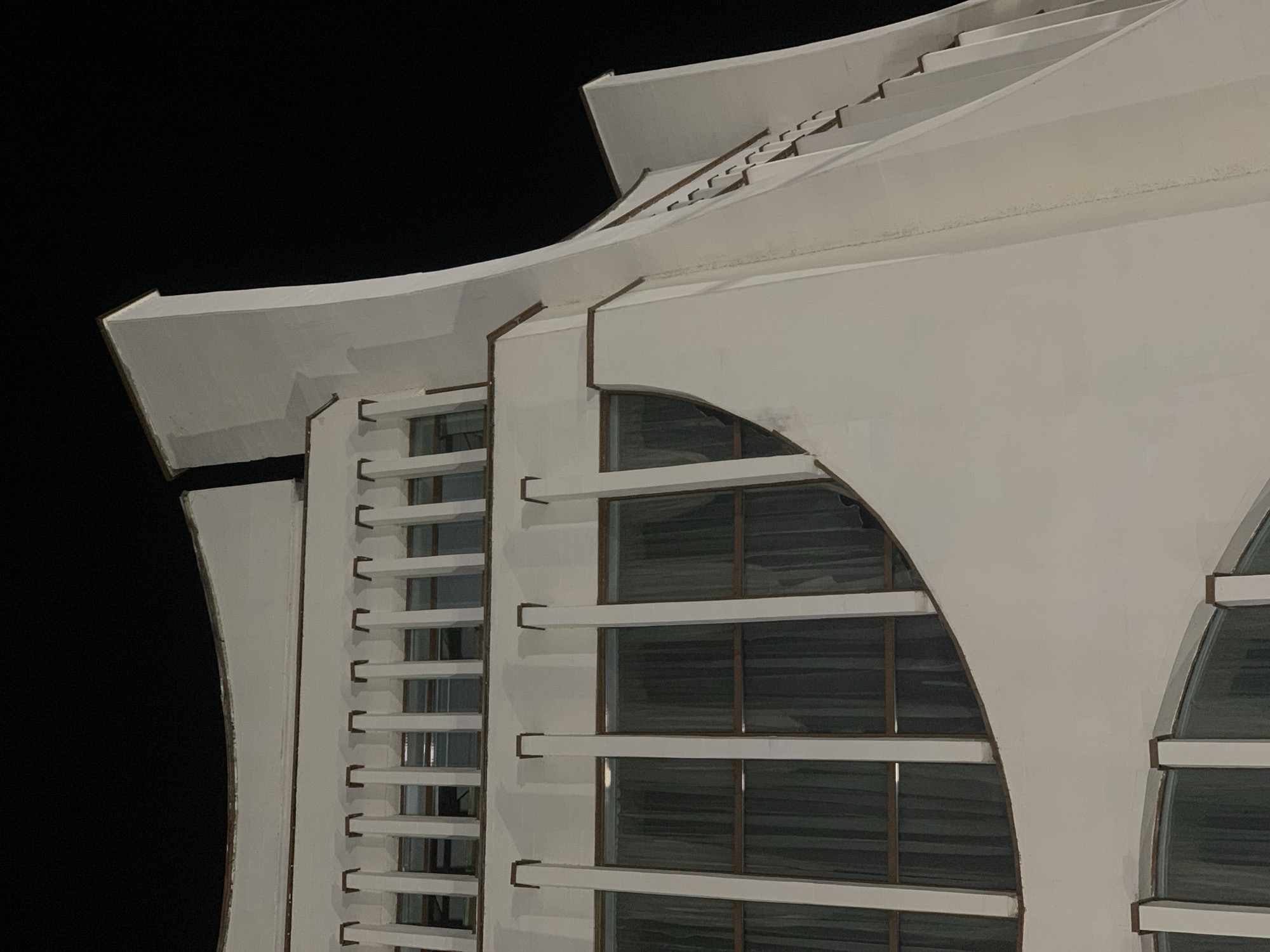 Here is a detailed description of the image:
The image depicts an architectural detail of a building, specifically the Drama Theatre in Hrodna, Belarus, at night. The building is predominantly white with dark accents. The architectural style features layered, projecting roof elements, creating a stepped effect toward the top. The lower portion of the building visible in the frame is characterized by vertical, slat-like structures and large, arched window openings with horizontal dividers. The windows are dark, reflecting the night. The overall impression is of a modern or contemporary architectural design. The background is a solid black, suggesting nighttime conditions and isolating the building as the primary subject.