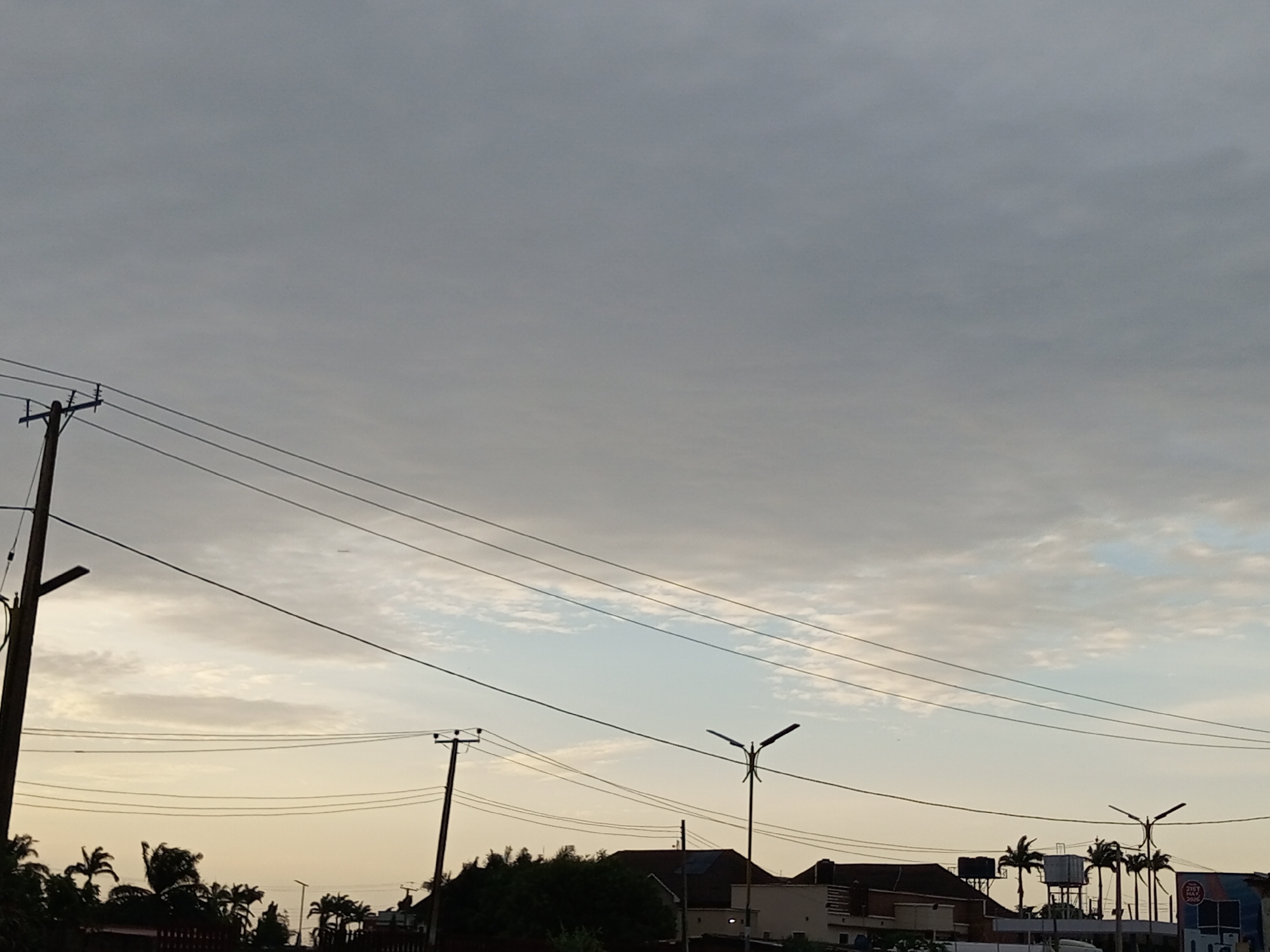 The image depicts a cloudy sky above a street scene in Barden Yana, Nigeria. Two telephone poles with wires are visible, one on the left and another on the right side of the frame. In the background, there are buildings, palm trees, and streetlights. The sky is overcast with a mix of gray and lighter clouds, with hints of pale blue. The overall lighting suggests it is either early morning or late afternoon. There are no people or vehicles prominently featured in the image.