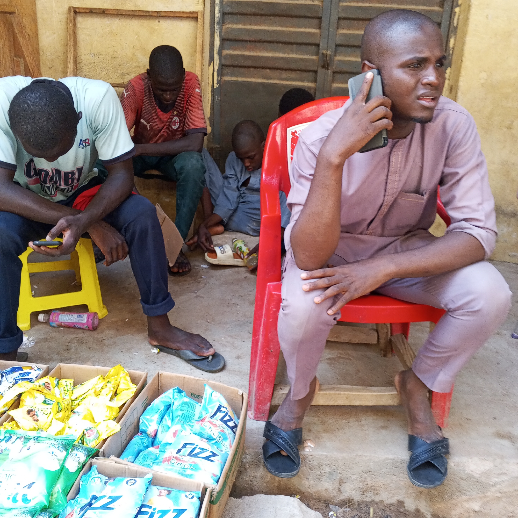 The image depicts a street scene in Gombe, Nigeria, with several individuals present. In the foreground, a man in a light brown garment is seated on a red plastic chair, engaged in a phone conversation. He is holding a dark-colored mobile phone to his right ear. To his left, another man in a light-colored t-shirt and dark trousers is seated on a yellow plastic stool, looking down and holding a small black object, possibly a phone, in his left hand. In the background, two other individuals are partially visible, one wearing a red t-shirt and dark trousers, seated on the ground, and another, a younger person, also seated on the ground in a gray garment. Several cardboard boxes filled with packaged goods, some with yellow and blue packaging, are arranged on the ground in the foreground. A pink bottle is visible near the yellow stool. The environment appears to be an outdoor area with a building structure in the background and a dirt ground surface.