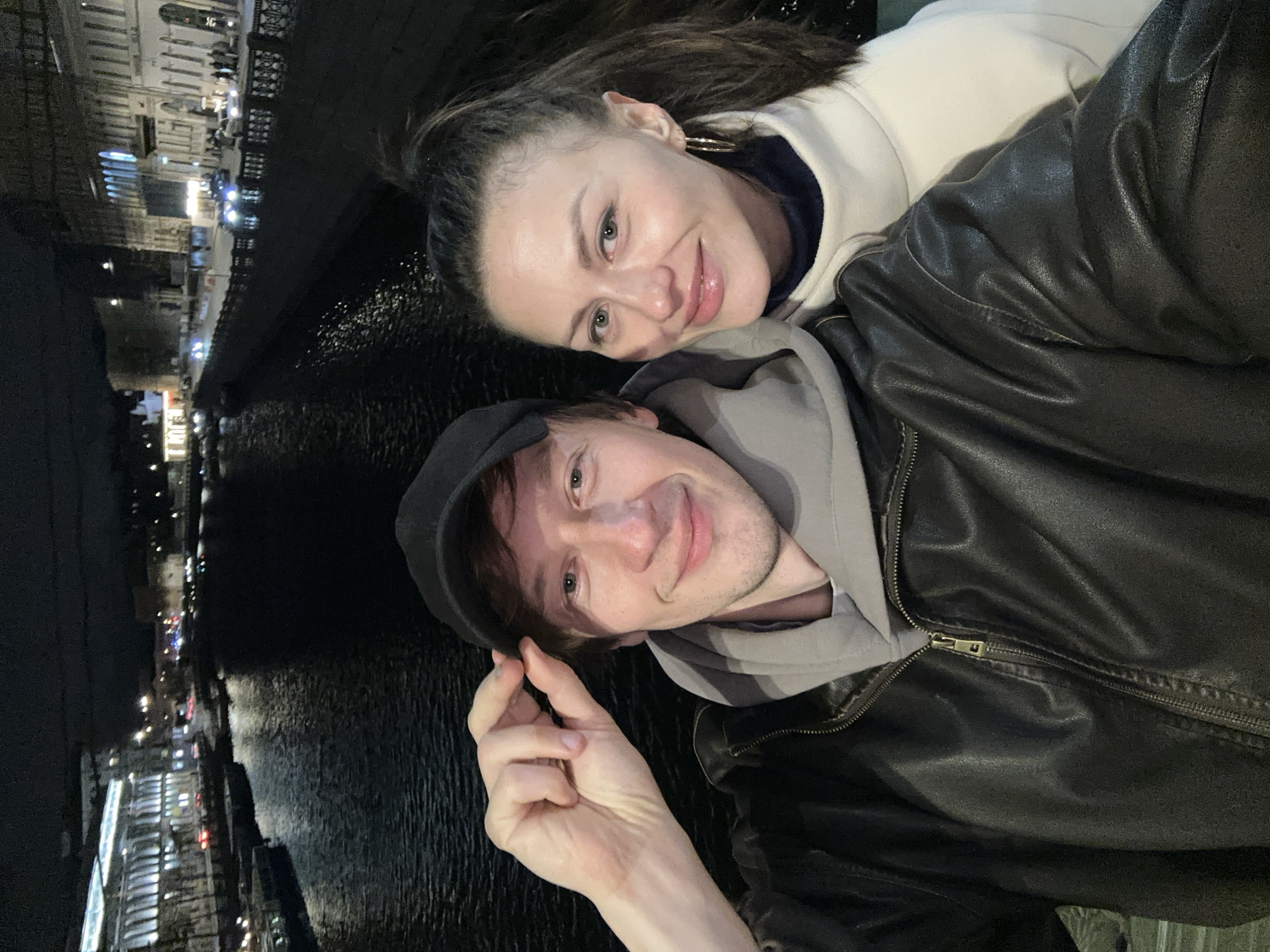 Two people are posing for a selfie outdoors at night in Saint Petersburg, Russia. The woman has dark hair, fair skin, and is wearing hoop earrings and a white sweater with a dark collar. She is smiling slightly. The man is wearing a black cap, grey hoodie, and a black leather jacket. He is also smiling and holding his fingers in a pinching gesture. The background shows a dark body of water, possibly a canal or river, reflecting lights from buildings and street lamps. The cityscape along the waterfront is illuminated.