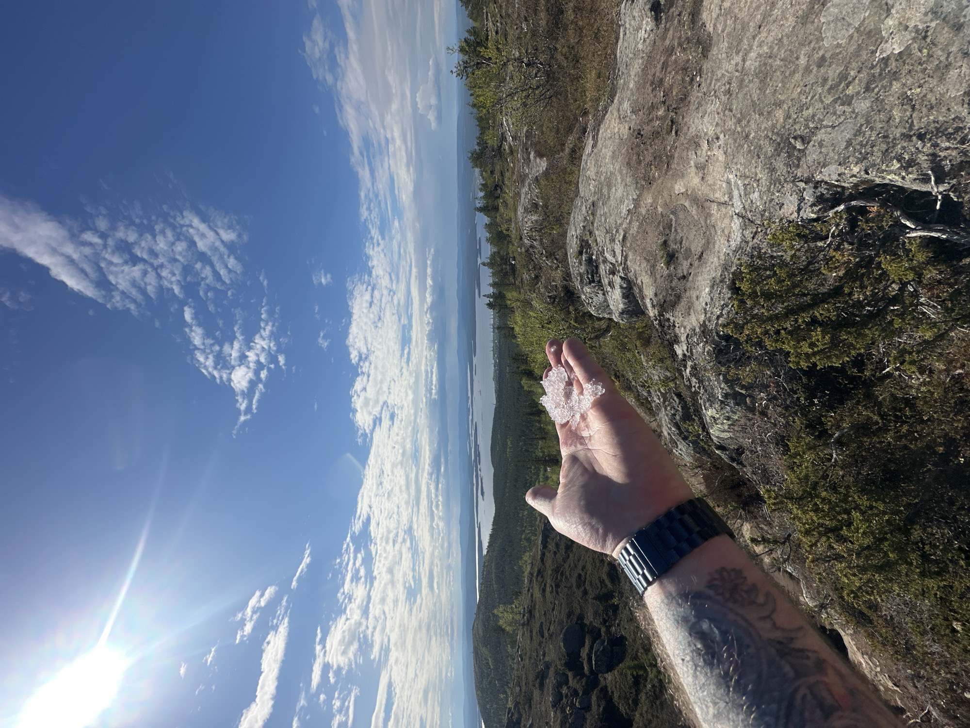 The image shows a man's arm and hand holding some small chunks of ice or frozen material. The person is wearing a black wristwatch on their left wrist. The arm is tattooed. In the background, there is a landscape featuring a body of water and wooded hills or mountains under a blue sky with scattered clouds. The scene may be from around Kandalaksha, Russia, a city located on the shore of the White Sea in the Murmansk Oblast. The environment suggests a natural, possibly elevated location overlooking the water and surrounding terrain.
