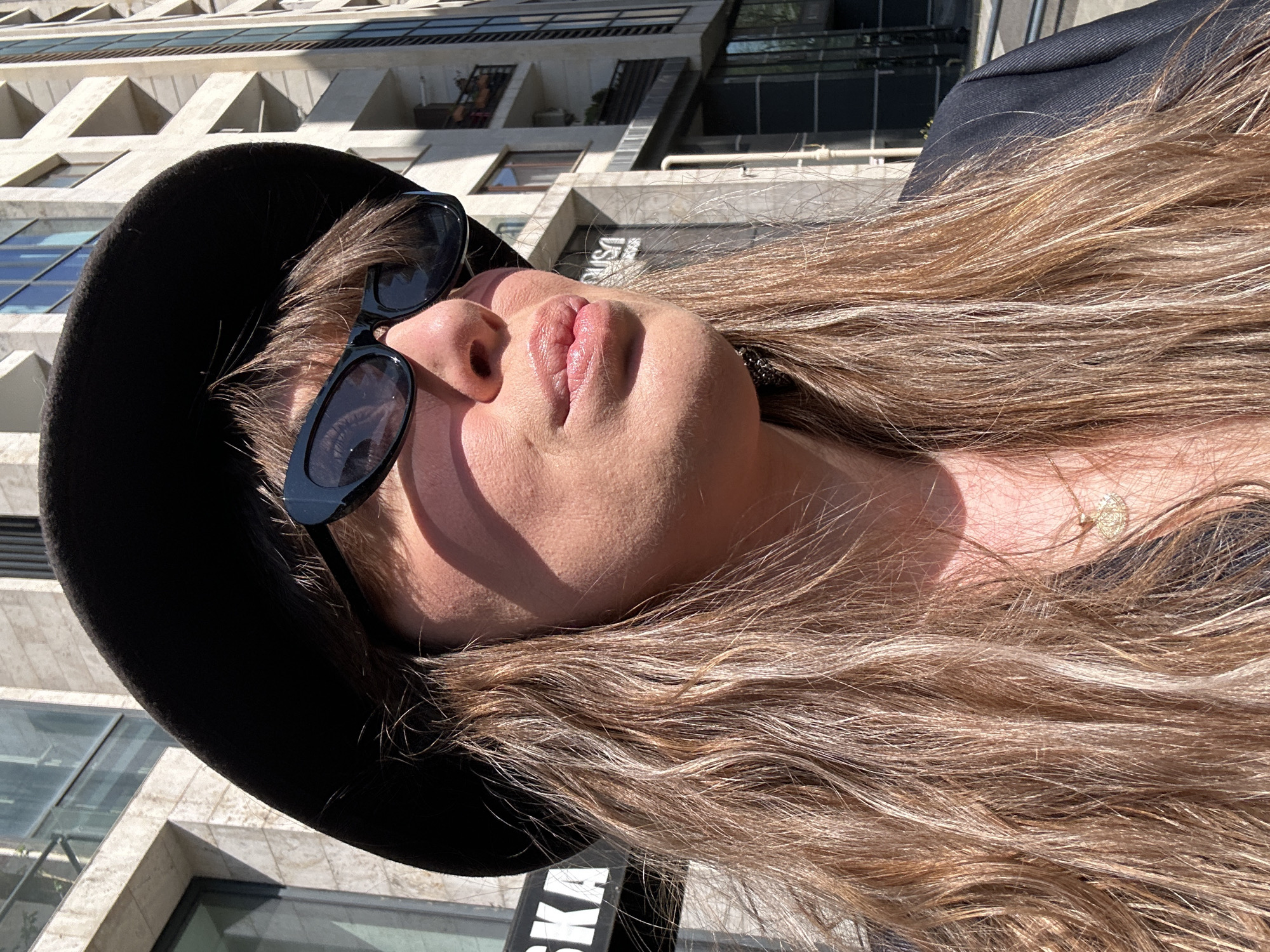 A close-up, slightly rotated portrait captures a woman outdoors in an urban setting, likely Tbilisi, Georgia, on a bright, sunny day. The subject, seen from the chest up, has long, wavy brown hair with lighter streaks, which flows down and across the frame. She wears a black fedora-style hat and dark, rectangular sunglasses, positioned on her face. A thin, delicate gold necklace with a small circular pendant is visible on her neck. Her head is tilted slightly back, and she appears to be looking upwards, with direct sunlight creating distinct shadows on her cheek and under her chin, highlighting the contours of her face and lips.
In the background, a striking modern building with concrete facades and geometric, angular balconies or protrusions is visible. Its multi-story structure features numerous windows, some reflecting the blue sky. The architecture suggests a contemporary design, possibly influenced by Brutalism. Part of a dark sign with white, uppercase, inverted text "USKA" (or similar) is discernible beneath her hair and shoulder. The clear skies and strong shadows indicate it is midday or early afternoon. The overall scene conveys a casual, relaxed moment in a vibrant city environment.