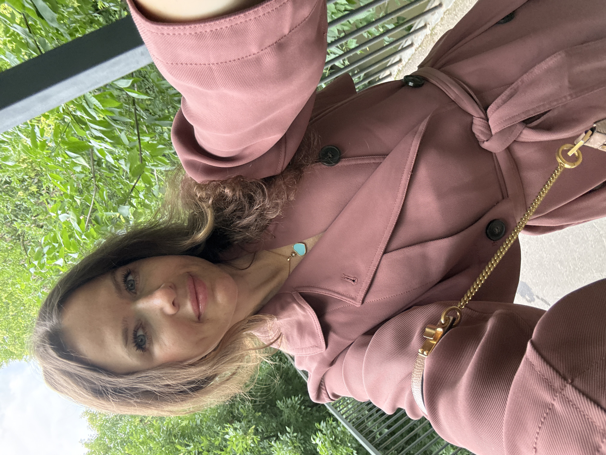 A woman with shoulder-length brown hair is taking a selfie outdoors. She is wearing a rose-colored trench coat cinched at the waist with a matching belt. A gold chain with a small purse is attached to the coat's belt. She has a light blue heart-shaped pendant necklace visible. The background consists of lush green foliage and a black metal railing. The setting appears to be a park or garden area within a city. There is no indication that this location is in Moscow, Russia.