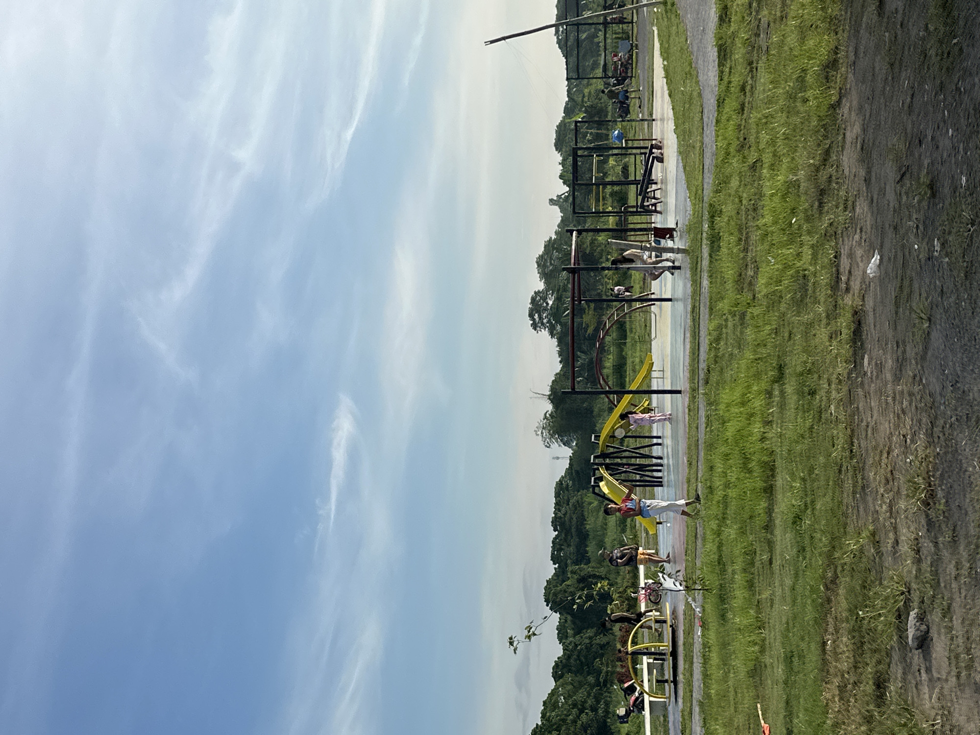 The image depicts an outdoor exercise park in Davao City, Philippines, featuring several individuals engaging in physical activities. There are exercise stations, a playground with slides, and grassy areas. People are using the exercise equipment and interacting within the park setting. The environment includes green spaces, trees, and infrastructure associated with a public recreational area. The sky is visible, suggesting a daytime setting.