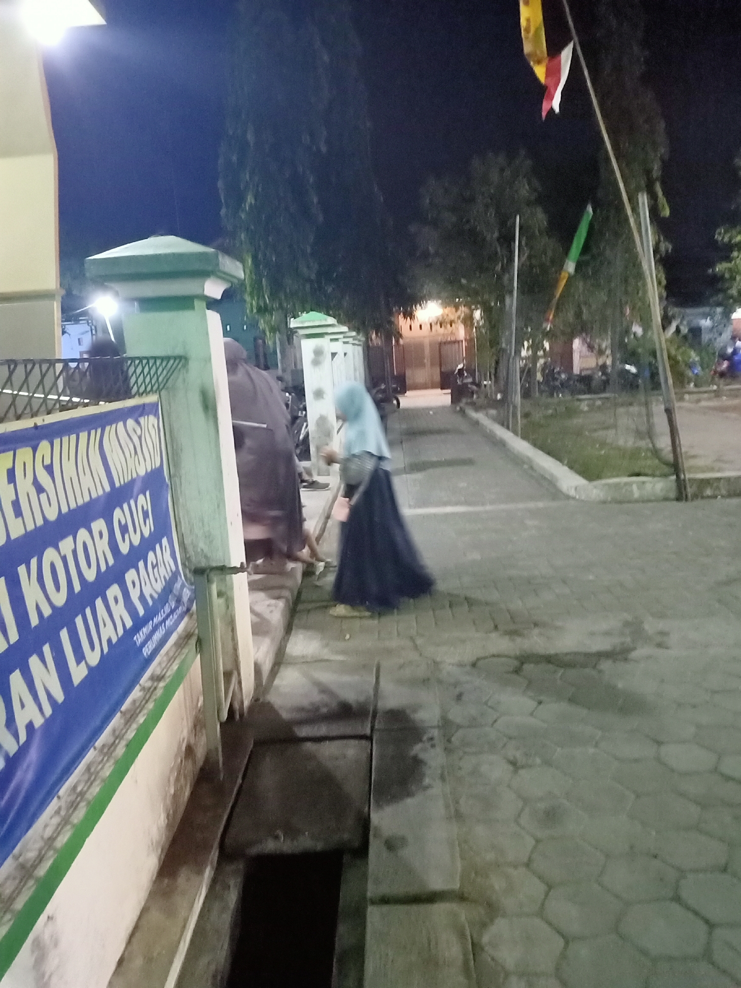 A medium shot captures an outdoor scene at night, illuminated by artificial lighting. In the foreground on the left, a blue banner with white lettering, partially visible, reads "BERSIHAK MA..." and "I KOTOR CUCI" and "AN LUAR PAGAR". To the right of the banner, a person wearing a light blue hijab and a dark, flowing skirt is sweeping the tiled ground with a broom. Behind this person, a dark-colored figure is partially visible, appearing to be squatting. A green-topped pillar is prominent to the left of the banner, and further back, trees and other vegetation are silhouetted against the dark sky. Flags, including one with red and white elements, are visible in the upper right portion of the frame, attached to poles. The ground is paved with interlocking hexagonal tiles. A small, dark-colored ditch or drain is visible in the lower center of the image. The overall impression is of a public outdoor area, possibly near a place of worship or community building, undergoing cleaning activities.