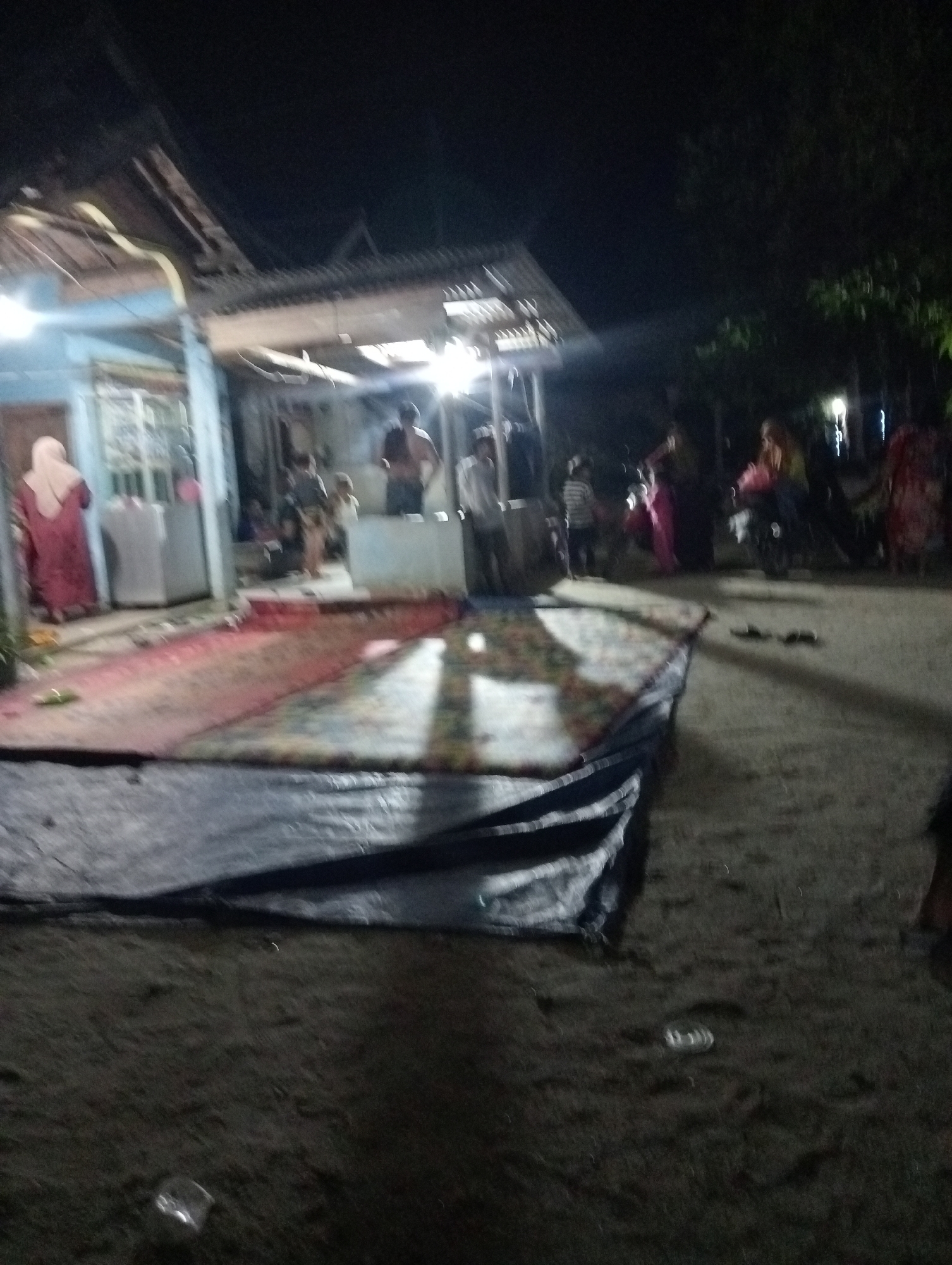 In the dimly lit outdoor area of Rasuan Baru, Indonesia, a group of people are gathered near a simple structure with a corrugated metal roof. A woman in a red hijab and dress stands to the left, facing away from the camera. In the center, a person appears to be shirtless and is looking towards the right. Several other individuals, including children, are present in the background. A large, patterned rug is spread out on the ground, partially covered by a dark tarp. The ground appears to be dirt or sand. The scene suggests a communal gathering or event taking place in a residential area at night, with artificial lighting casting shadows.