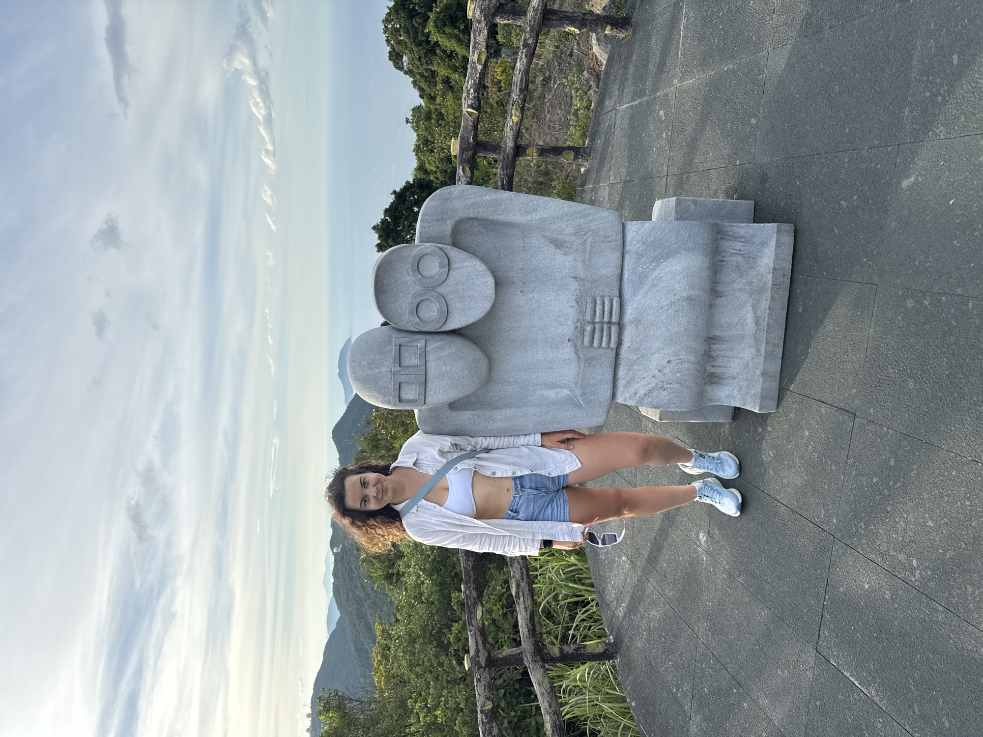 A young woman with curly brown hair stands next to a large, gray stone sculpture of two abstract figures. She is wearing a white crop top, a white open button-down shirt, and denim shorts. She has light blue sneakers and is holding a pair of white-rimmed sunglasses in her right hand. The sculpture is positioned to her right, and depicts two figures with block-like heads and circular eye shapes. They are in a close embrace, with their arms interlocked.
The woman and sculpture are on a paved platform with dark gray square tiles. A wooden railing with weathered posts and crossbars runs along the left and right edges of the platform. Behind the railing, a lush green hillside slopes downwards, covered in dense trees and foliage. In the distance, a vast expanse of blue ocean stretches to the horizon, meeting a sky filled with scattered, wispy clouds. The overall setting appears to be a scenic overlook, likely in the Sơn Trà Peninsula region of Đà Nẵng, Vietnam, given the mountainous terrain and coastal views characteristic of the area. The light suggests it is either early morning or late afternoon.