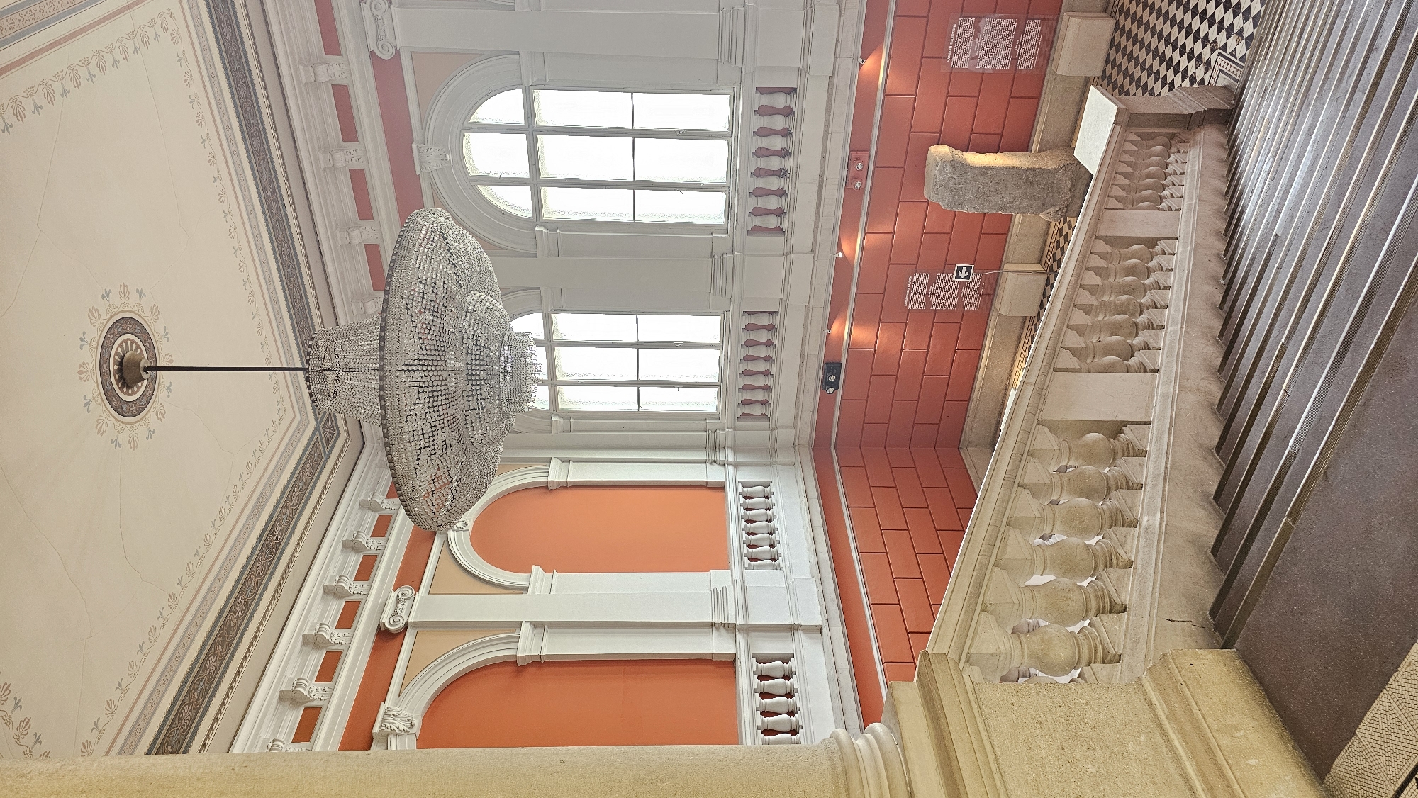 The image shows an interior view of the Varna Archaeological Museum in Bulgaria, specifically the main staircase and the ceiling above. The ceiling is high and ornate, featuring a large crystal chandelier, decorative moldings, and painted patterns. Two arched windows provide natural light. The walls are painted in shades of orange and peach, and there are classical-style columns and architectural details. To the right is the staircase, constructed with stone and metal railings. The stairs lead to a checkered black and white tiled floor. A stone artifact sits near the staircase, and informational placards are mounted on the walls.