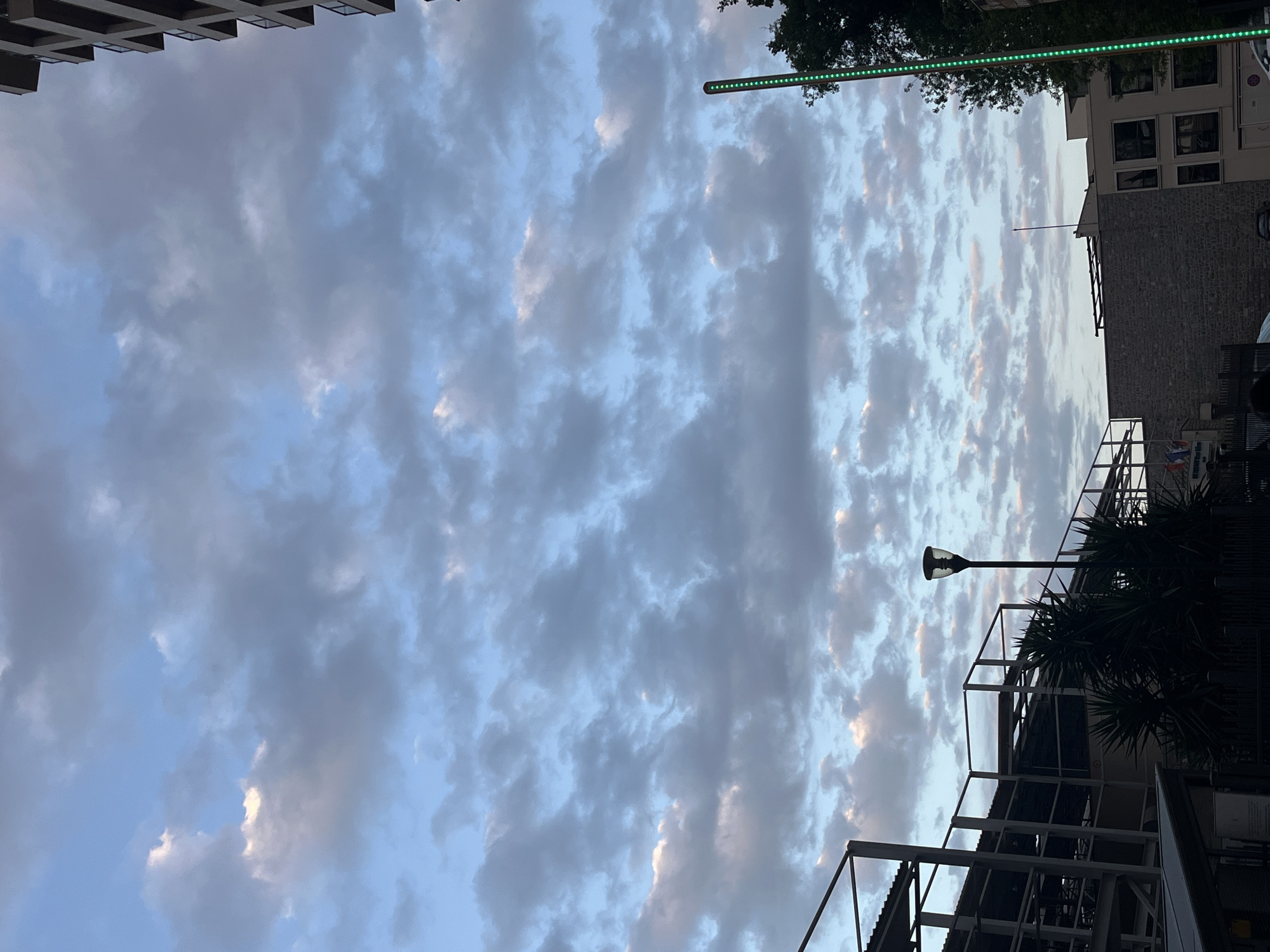 The image depicts a view looking up at the sky in Boreti, Montenegro. The sky is filled with clouds, illuminated with muted oranges. At the top of the frame is the top of a building. On the right is a building façade, a green LED strip light, street lighting, a multi-level metal structure, and greenery.
