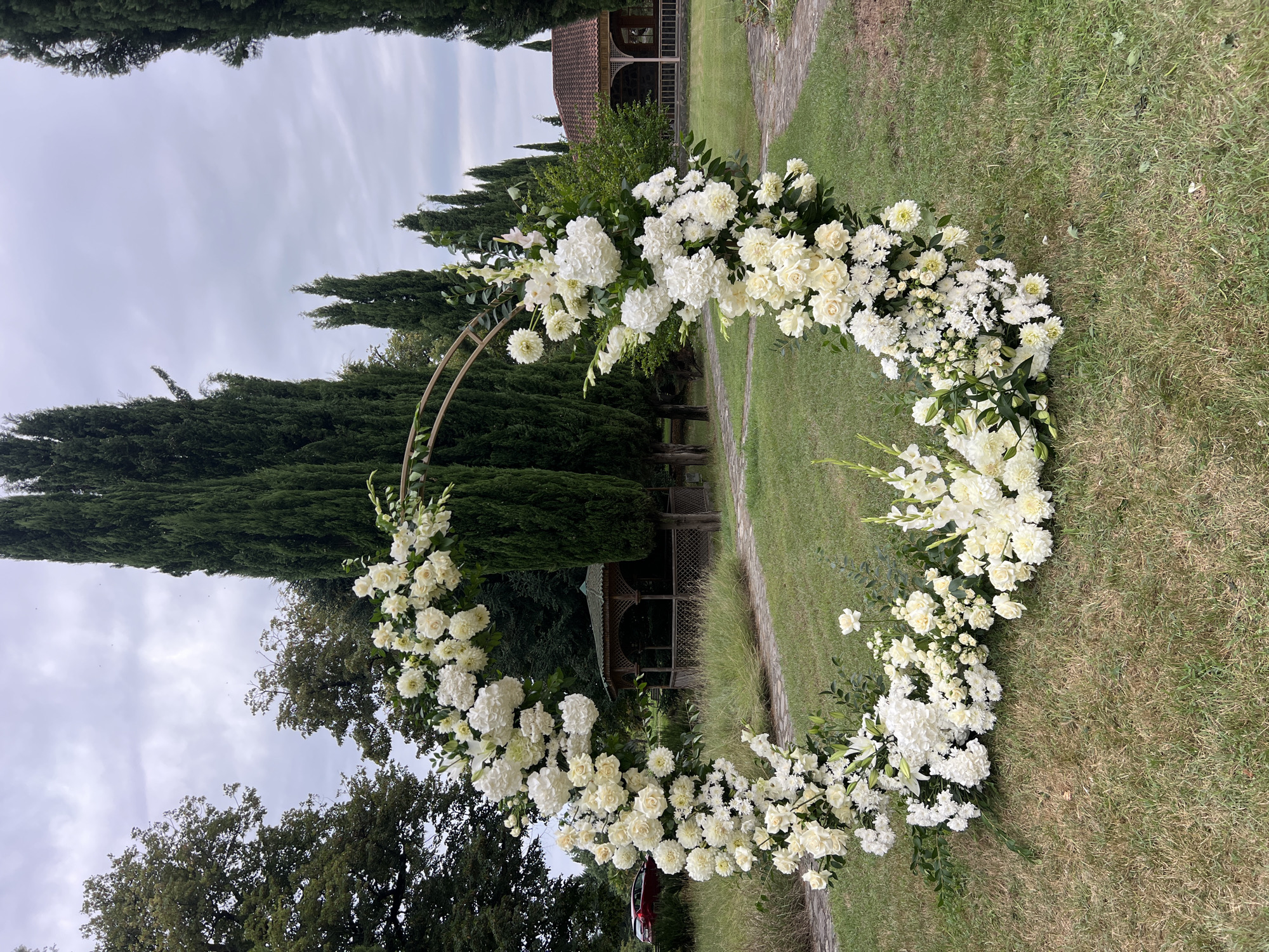 The image shows a circular floral archway, predominantly adorned with white roses and hydrangeas, set on a grassy lawn. The archway is positioned outdoors, with a backdrop of tall cypress trees and lush green foliage. In the middle ground, a wooden gazebo and a building with a tiled roof are visible. A red car is parked to the left of the scene. The sky is overcast. The location is identified as Napareuli, Georgia.