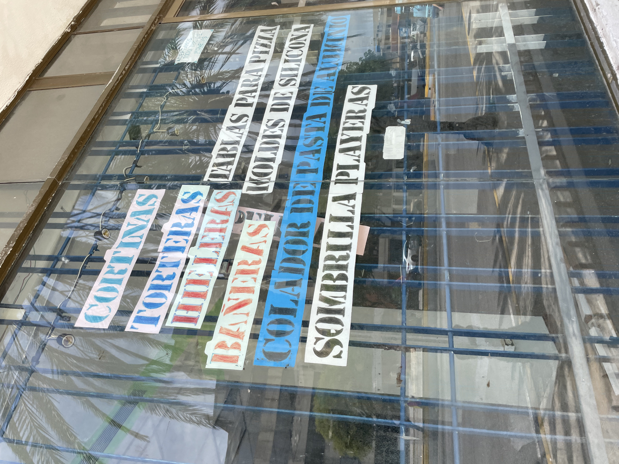The image shows a storefront window in Porlamar, Venezuela, with various handwritten signs advertising products. The signs, some on colored paper, are taped to the glass and list items such as "CORTINAS" (curtains), "TORTESAS" (cakes), "HIELERAS" (ice boxes), "BANERAS" (baths), "COLADOR DE PASTA" (pasta strainer), "MOLDES DE SILICONA" (silicone molds), "SOMBRILLAS PLAYERAS" (beach umbrellas), and "TABLAS PARA PIZZA" (pizza boards). The window is covered by a blue metal grate, and palm tree fronds are visible reflected in the glass, indicating a tropical or subtropical location. Outside the window, a street scene with vehicles and buildings can be seen, suggesting an urban environment. The lighting indicates it is daytime.