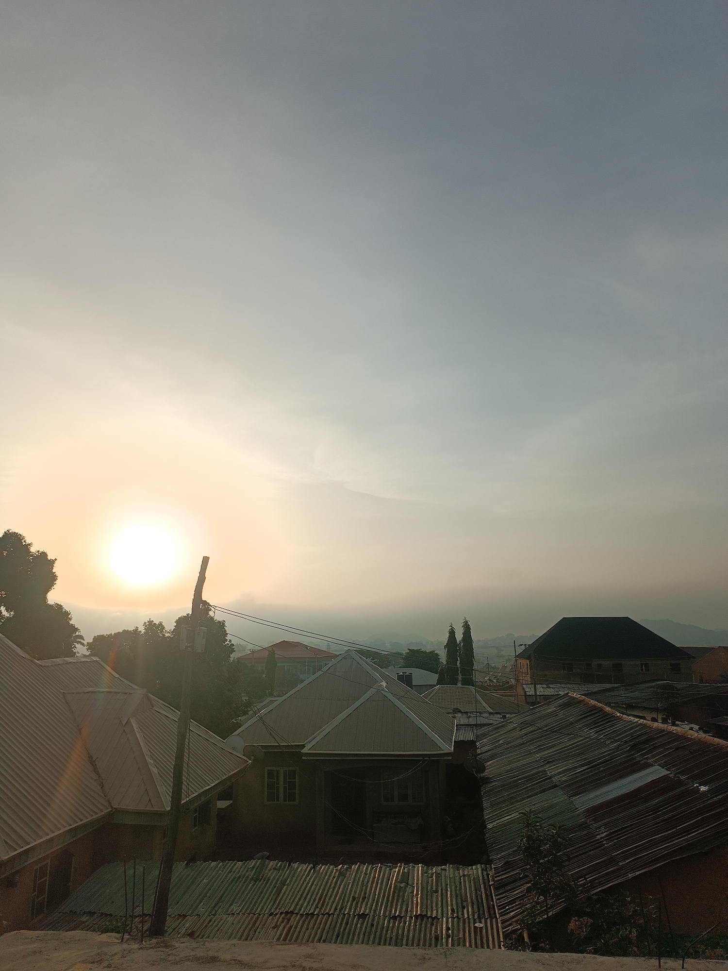 The image is a ground-level, outdoor shot of a cityscape with a bright, diffuse sun in the upper left. The sky is a pale, hazy mix of white and blue. The foreground is filled with rooftops made of corrugated metal. A utility pole with wires runs vertically near the left side of the frame. In the middle distance are trees and several buildings, including a dark-roofed structure on the right. The background shows misty hills. The location is Bukuru, Nigeria, as confirmed by image source. The scene captures a typical morning or late afternoon in an urban setting. There are no people visible.