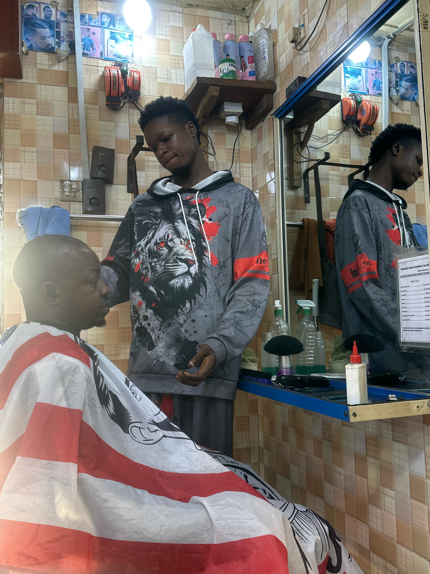 The image depicts the interior of a barbershop in Badagry, Nigeria. A barber, wearing a grey hooded sweatshirt with a lion graphic and red accents, is attending to a customer. The customer, with a shaved head, is seated and draped with a cape featuring red and white stripes and a black graphic. The barbershop environment is characterized by tiled walls in shades of beige and light brown, with various framed pictures of hairstyles displayed. Electrical outlets and two orange barber clippers are visible on the wall. A mirror reflects the scene, showing more framed pictures and a shelf with bottles of hair products and sprays. A blue shelf below the mirror holds barbering tools and supplies, including clippers, brushes, and bottles. A price list for haircuts is affixed to the mirror.