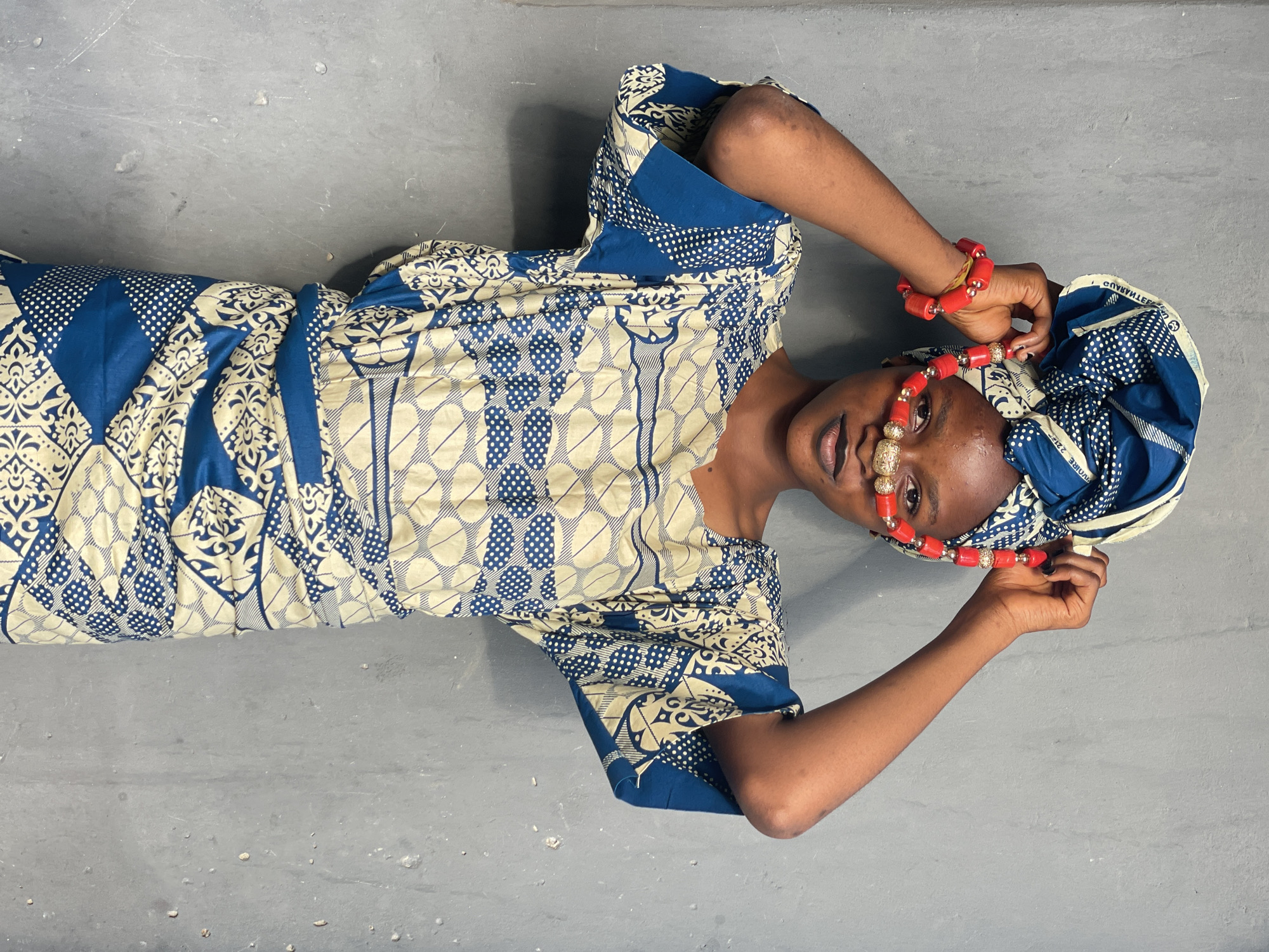 A woman is lying on a gray surface wearing a blue and beige patterned dress and headwrap. She is wearing red beaded jewelry on her head and wrists. Her hands are raised near her head. The image has been rotated. The location is not discernible from the given image.