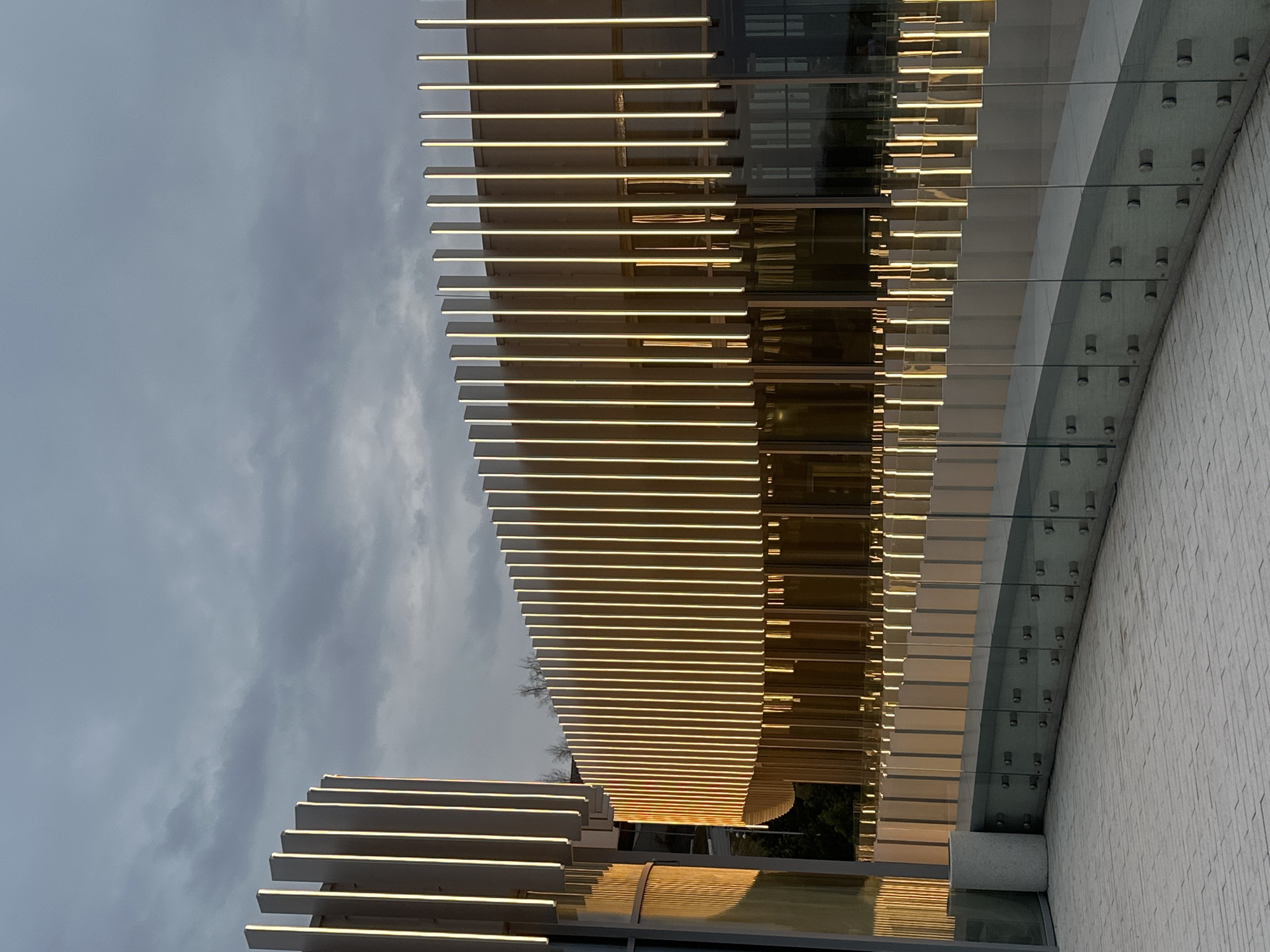 The image shows a section of modern architecture, likely a building in the city of Kazan, Russia. The building features a facade with horizontal illuminated slats, creating a layered effect. A glass and stone section is attached to the primary structure, secured by multiple visible bolts. The sky overhead is cloudy and overcast.