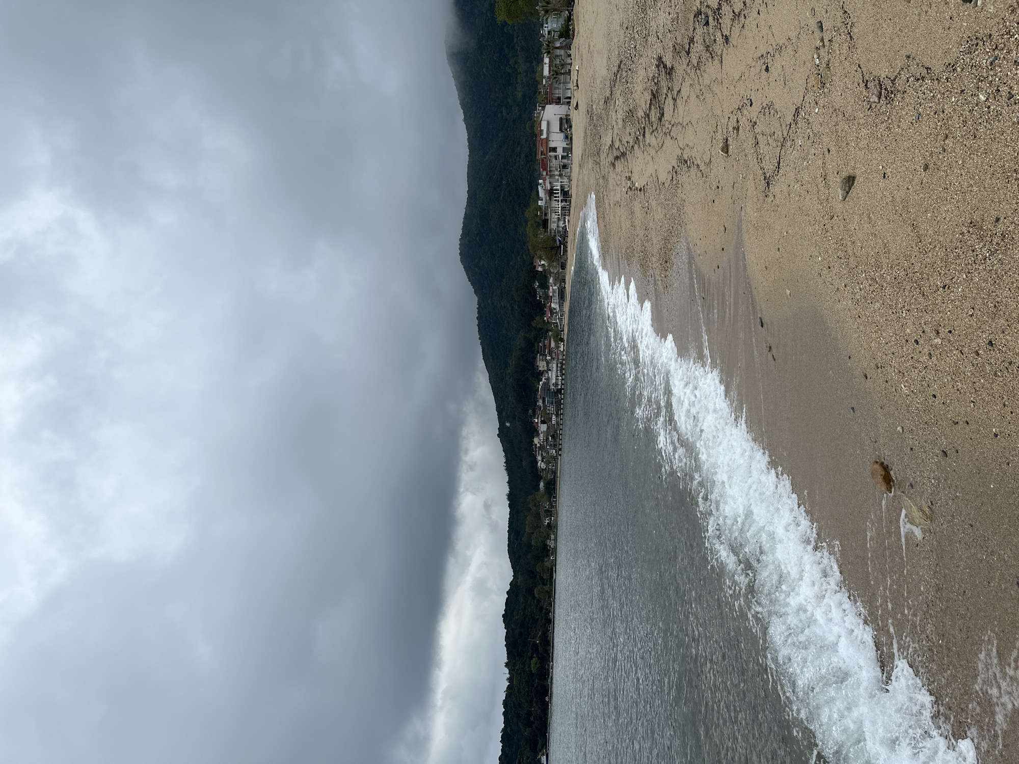 The image shows a beach scene in Ολυμπιάδα, Greece. The shoreline is composed of sand and small pebbles. Waves are breaking along the shore. In the background, there is a mountain range covered in green trees. A few buildings are visible along the shoreline. The sky is overcast with grey clouds.