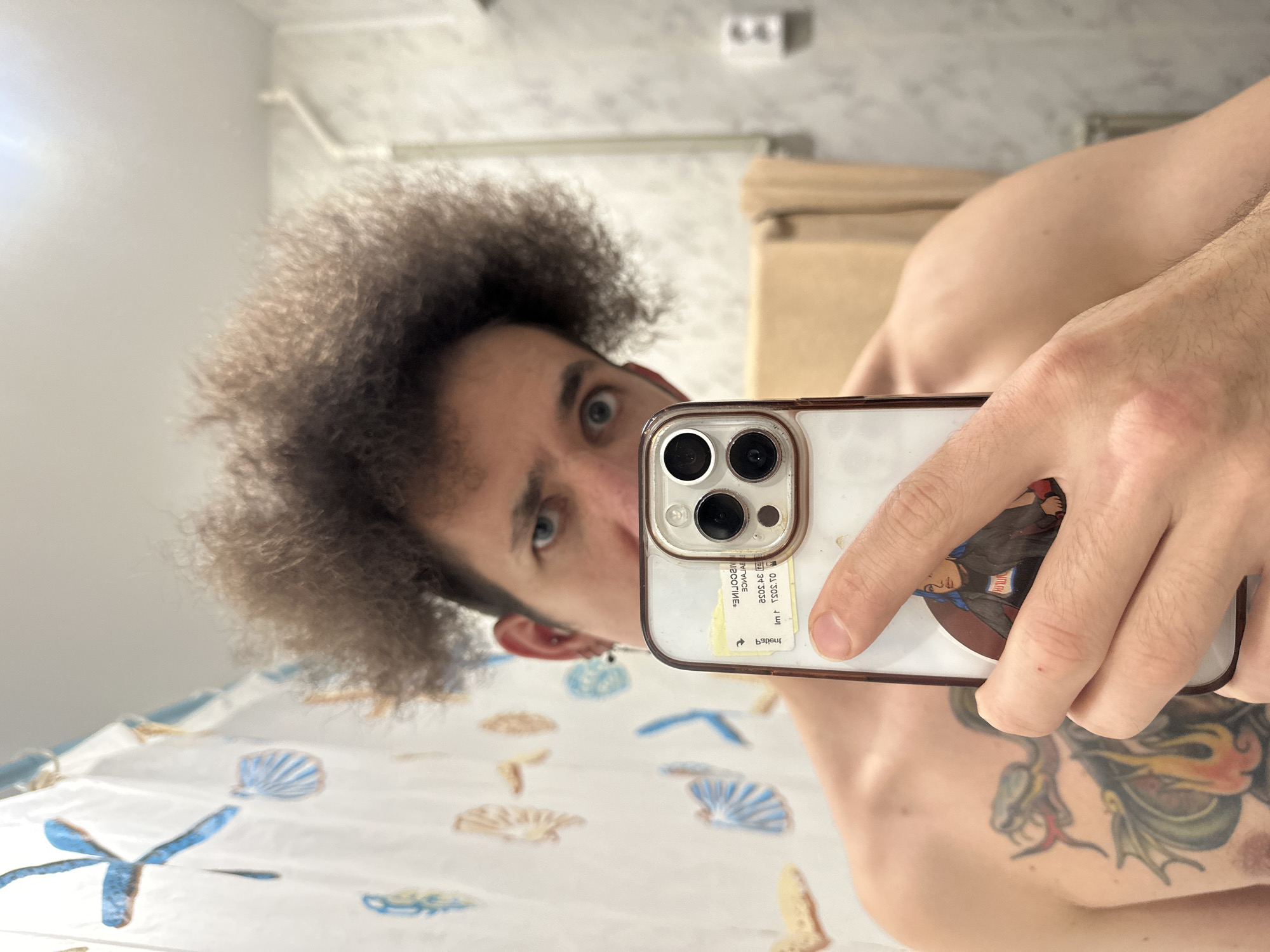 A selfie taken in a bathroom shows a person holding a smartphone with a clear case that has a sticker of a woman with blue hair. The person has an afro and is looking at the camera, with their left ear visible. They are shirtless, revealing a tattoo on their chest of a dragon with wings. The background includes a shower curtain with a shell pattern, a towel rack with a towel, and a light switch. There is no visual information to suggest this location is in Saratov, Russia.