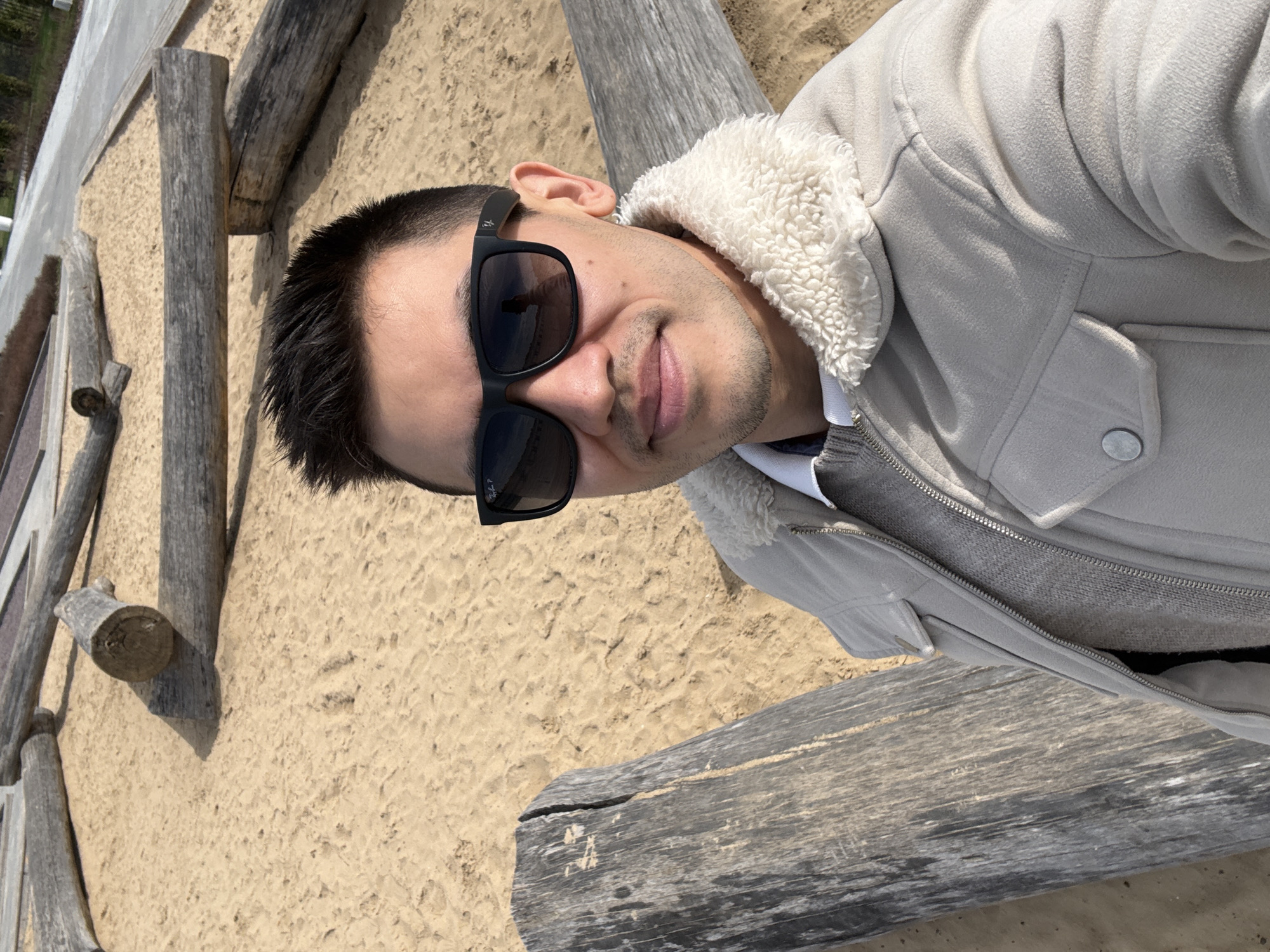 This image is a selfie of a young man, captured outdoors in Kazan, Russia, rotated approximately 90 degrees to the left. The man is shown from the chest up, looking directly at the camera with a subtle, relaxed smile. He has short dark hair and is wearing black rectangular sunglasses that reflect a bright, sunlit environment. A small, unidentifiable logo or text is visible on the temple of his sunglasses.
He is dressed in a light-colored, possibly beige or light grey, jacket featuring a prominent, fluffy white or cream-colored sherpa collar and inner lining. Beneath the jacket, a grey knitted sweater or similar garment is visible, along with a white shirt collar. The jacket has a metallic button on a flap on the right side and a zipper closure.
The background consists of light-colored sandy ground, suggesting a beach or a sandy pathway. Several weathered, rough-hewn wooden logs or beams are scattered across the sand, some appearing to be partially buried. The bright lighting indicates a sunny daytime setting, with distinct shadows cast by the wooden elements. The overall scene conveys a relaxed outdoor mood.