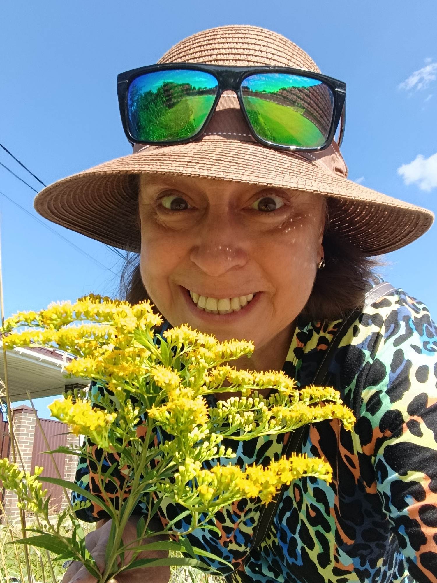 An outdoor, close-up, selfie-style photograph shows a woman wearing a wide-brimmed straw hat and sunglasses, holding a bouquet of small yellow wildflowers. The woman's face is visible from just below the sunglasses, and she has a wide, toothy smile and her eyes are looking directly at the camera. The sunglasses reflect a landscape of green fields and trees under a clear blue sky. The woman is wearing a colorful, leopard-print garment. Behind her, a blue sky with a few scattered clouds is visible, along with some power lines and a partial view of a building with a brown brick wall and a light-colored roof. The lower part of the image shows more of the yellow wildflowers and green foliage. The scene suggests a sunny day in a rural or semi-rural setting. The location is stated to be in the city of Vereya, Russia.