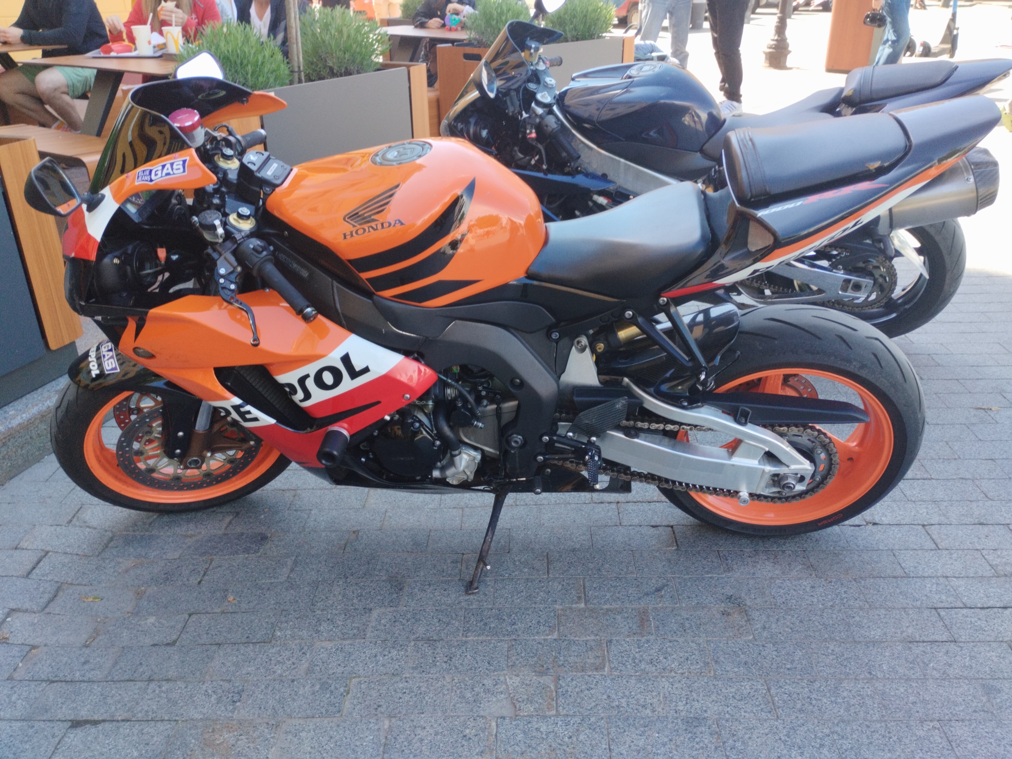 An orange and white Honda CBR1000RR motorcycle with Repsol branding is parked on a grey brick-paved surface. The motorcycle is angled towards the left, with its side stand deployed. Behind it, partially obscured, is a dark blue or black motorcycle of similar type. People are seated at tables in the background, some under umbrellas, likely at a cafe or restaurant. The presence of greenery in planters behind the tables suggests an outdoor dining area. This photo was taken in Odesa, Ukraine.