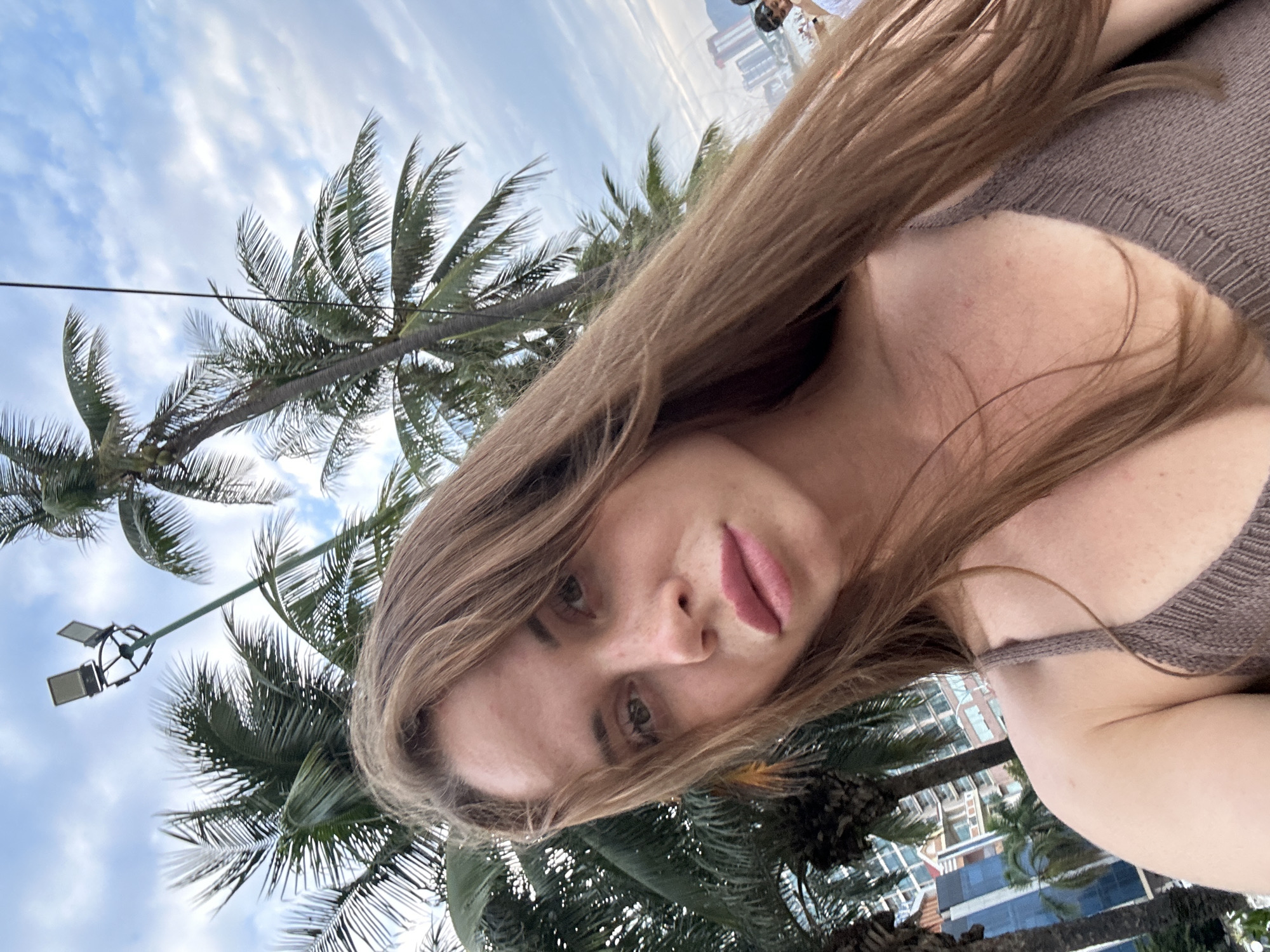 This vertical full-frame selfie-style portrait depicts a young woman outdoors, with the image rotated 90 degrees clockwise. The woman, who is the primary subject, has long, light brown hair cascading over her shoulders and chest. She has light skin, with visible freckles across her nose and cheeks, and her full lips are adorned with pink lipstick. Her eyes are light-colored, and she gazes directly at the camera with a neutral to slightly contemplative expression. She is wearing a light brown or beige knitted top, with a thin strap visible over her left shoulder.
The background is dominated by large, lush green palm trees, characteristic of a tropical environment. Between the palm fronds, a partly cloudy sky is visible, featuring light blue patches and scattered white clouds. In the distance, behind the dense foliage, modern high-rise buildings suggest an urban or resort setting. A distinctive green street light pole with multiple fixtures can also be seen in the upper left background.
The lighting indicates it is daytime, likely late morning or early afternoon, with soft, diffused sunlight under partly cloudy skies. The combination of palm trees and modern architecture strongly suggests a coastal resort city, consistent with the provided location context of Nha Trang Ward, Vietnam. No visible text is present in the image.