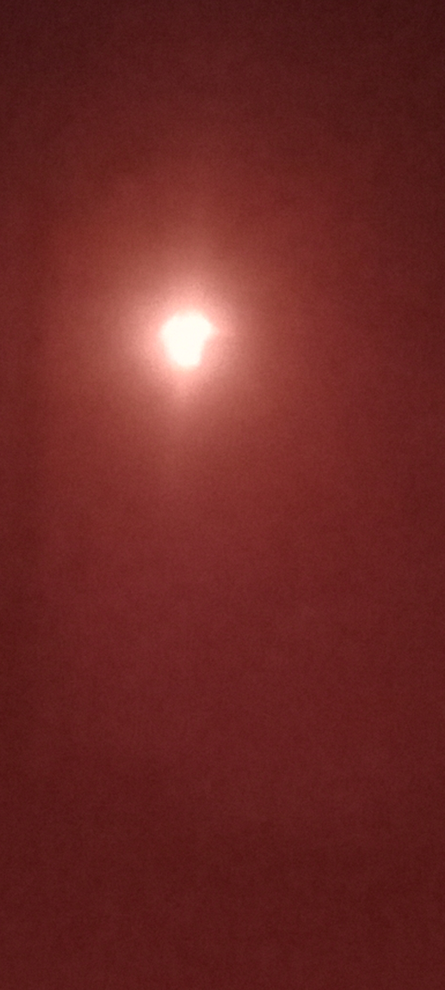 The image is a photograph of a red textured surface with a bright, irregular light source in the upper center. The light source appears to be an eclipse or a partial obscuration of a larger light. The overall color palette is dominated by shades of red. There are no discernible people, objects, or environmental features that would allow for the identification of a specific location, including Adai, Spain. The image is abstract and lacks detail to provide a forensic description.