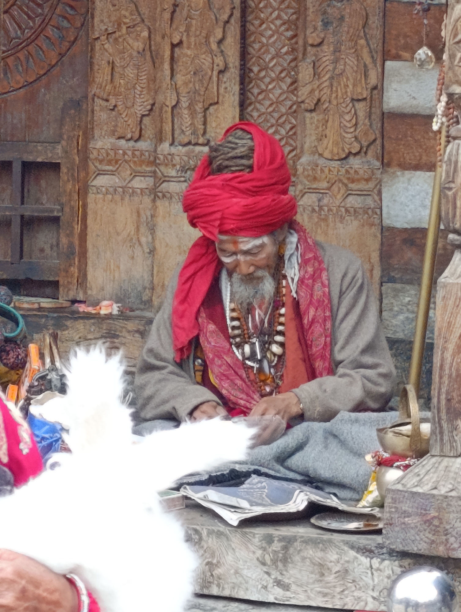 In Vashisht, India, a man with a gray beard and a red turban is seated on a wooden platform. He is wearing a gray robe, a red scarf, and multiple necklaces made of beads and shells. He is looking down and appears to be holding a white fluffy object in his hands, possibly engaged in a ritual or craft. The background consists of intricately carved wooden panels, suggesting a religious or cultural setting. Several objects are visible around him, including a metal pot, a golden staff, and what appear to be prayer beads. The overall scene is of a solemn and spiritual nature.