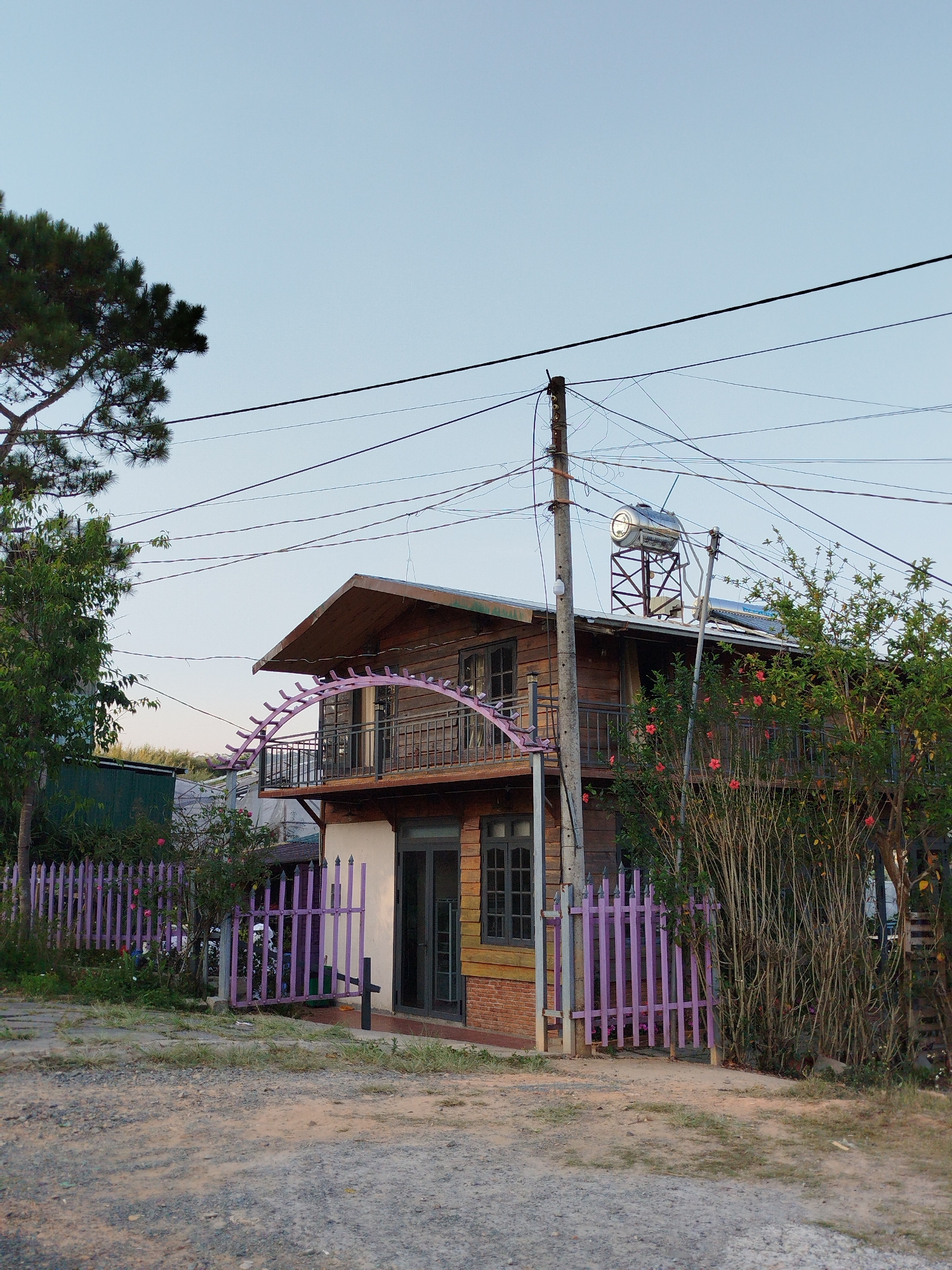 A two-story building stands in Da Lat, Vietnam. The building's upper story is wood paneling; the lower story is painted white and has a brick section. A purple fence surrounds the yard. A solar water heater sits on the roof. Overhead power lines are visible. A few trees and shrubs are in the yard. The ground is dirt and gravel.
