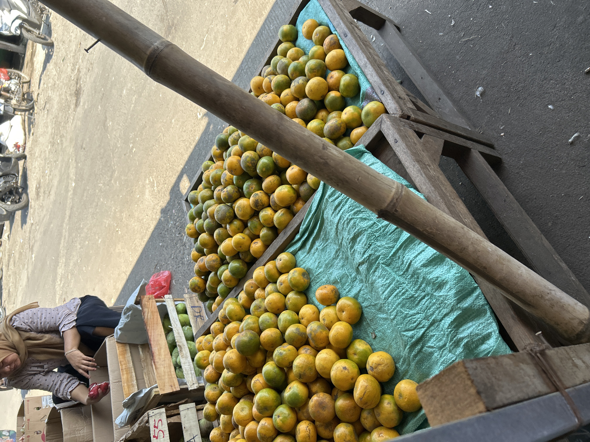 In a street market in Depok, Indonesia, a woman wearing a hijab and a patterned blouse crouches down, tending to a wooden crate filled with fruit. She is holding a red item in her right hand and appears to be sorting through it. In front of her are two wooden crates brimming with oranges, some green and some yellow, arranged on a teal-colored tarp. The crates are placed on wooden stands. In the background, a paved road is visible, with motorcycles parked along it, suggesting a bustling urban environment. A prominent bamboo pole runs diagonally across the foreground, partially obscuring the view of the market scene. The overall impression is that of a typical Indonesian street market, with an emphasis on fresh produce and daily commerce.