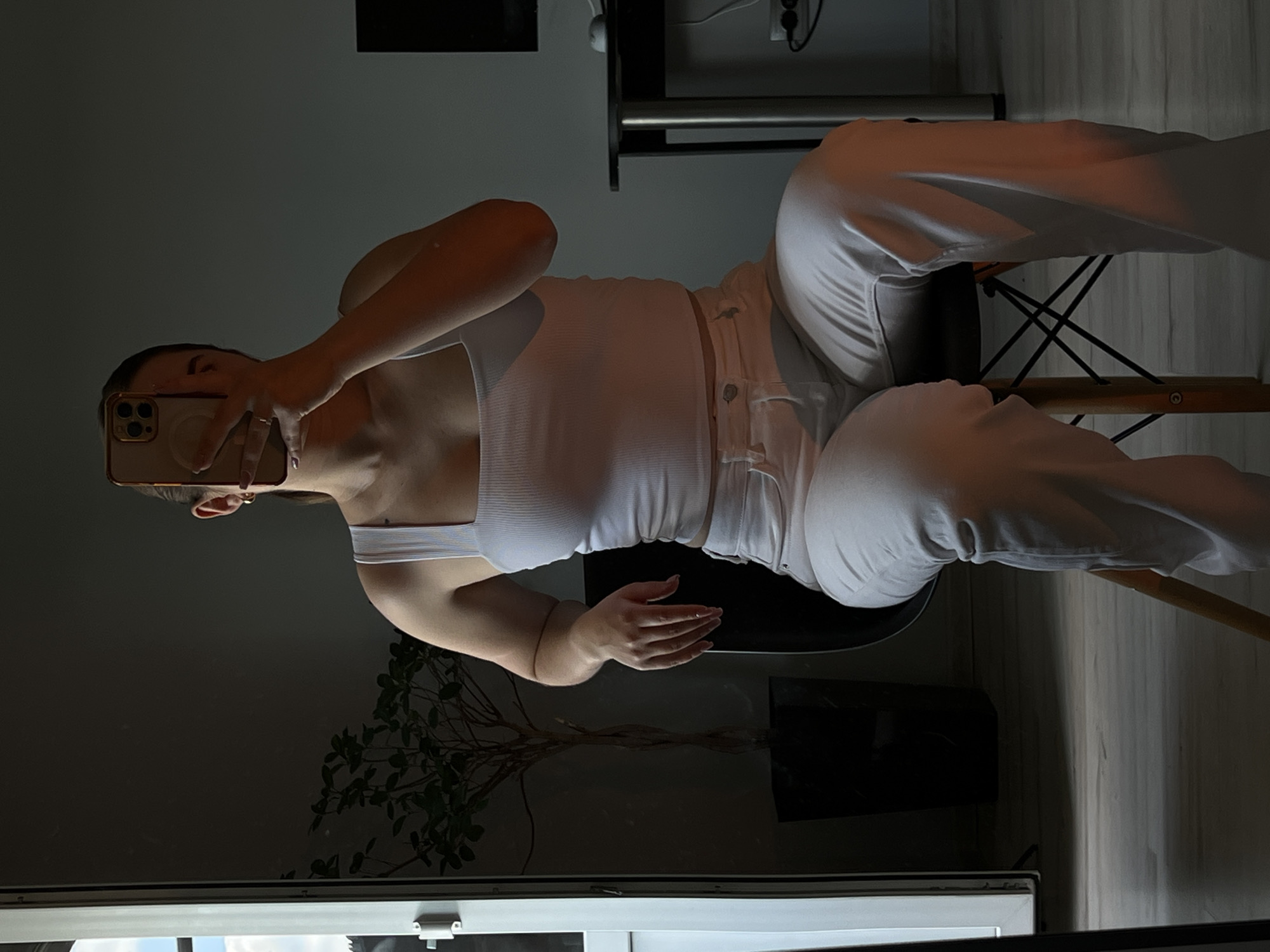 A young woman sits on a chair, taking a mirror selfie. She is wearing a white tank top and white pants. The background is a dimly lit room with a plant in a pot and a desk with a computer monitor. The woman is holding her phone up to her face, obscuring it. The lighting suggests it is either morning or evening. The image does not provide enough information to determine the specific location in Ternopil, Ukraine.