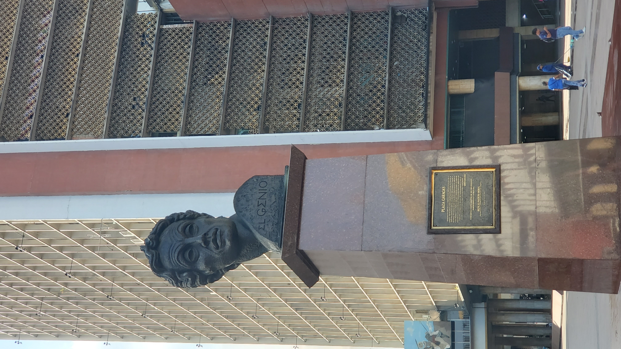 Here is a description of the image:
The image shows a bust of El Gizio (Greek for "The Genius") located in Plaza Caracas, Caracas, Venezuela. The bust is mounted on a tall, polished stone pedestal. A bronze plaque is affixed to the pedestal. Behind the bust is a building with latticed windows. People can be seen walking in the background.