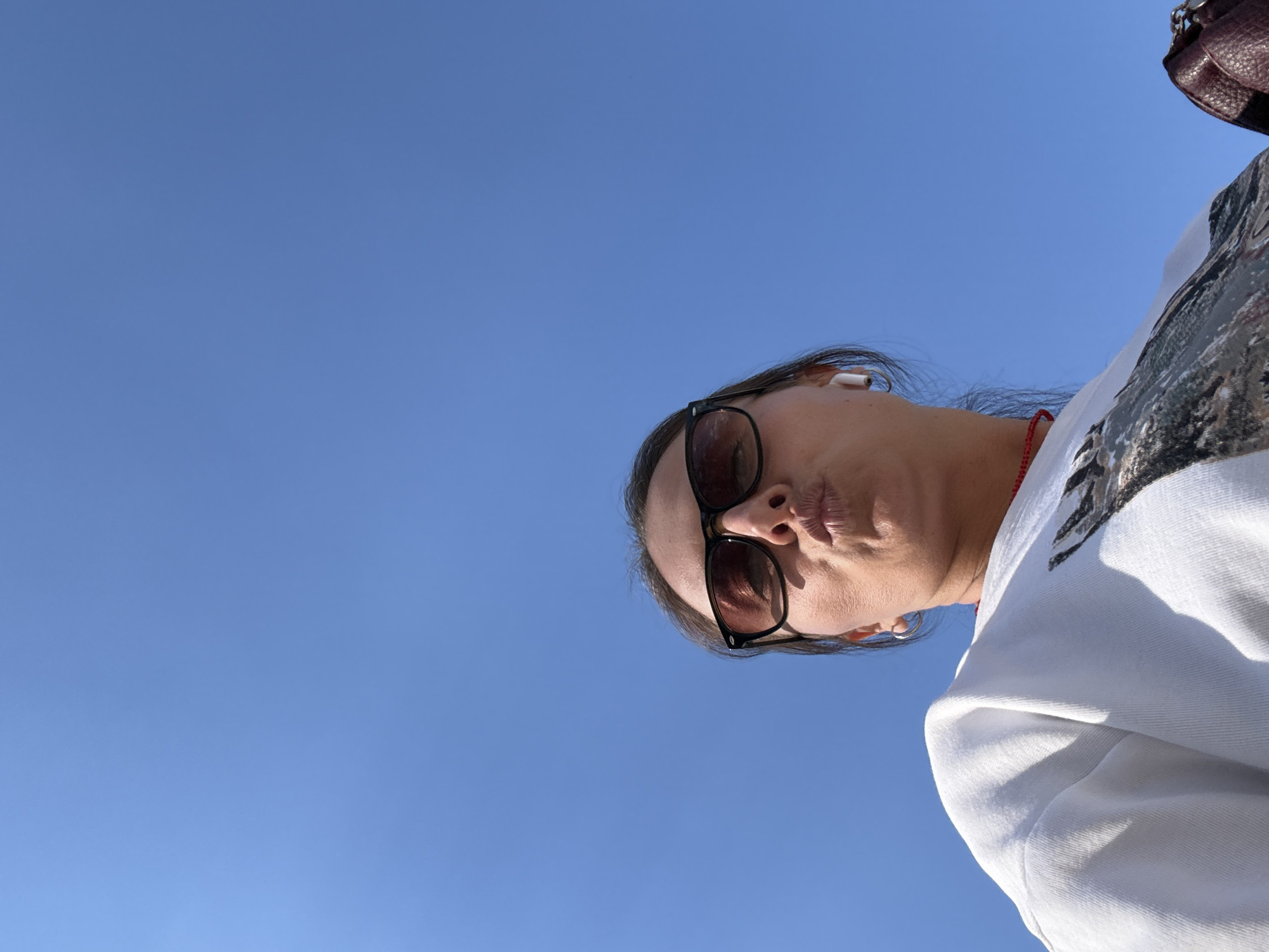 The image shows a woman in sunglasses and a white sweatshirt looking down at the camera. She has an earbud in her ear and a red necklace. Part of a handbag is visible in the upper right corner. The background is a clear blue sky. The location is not discernible.