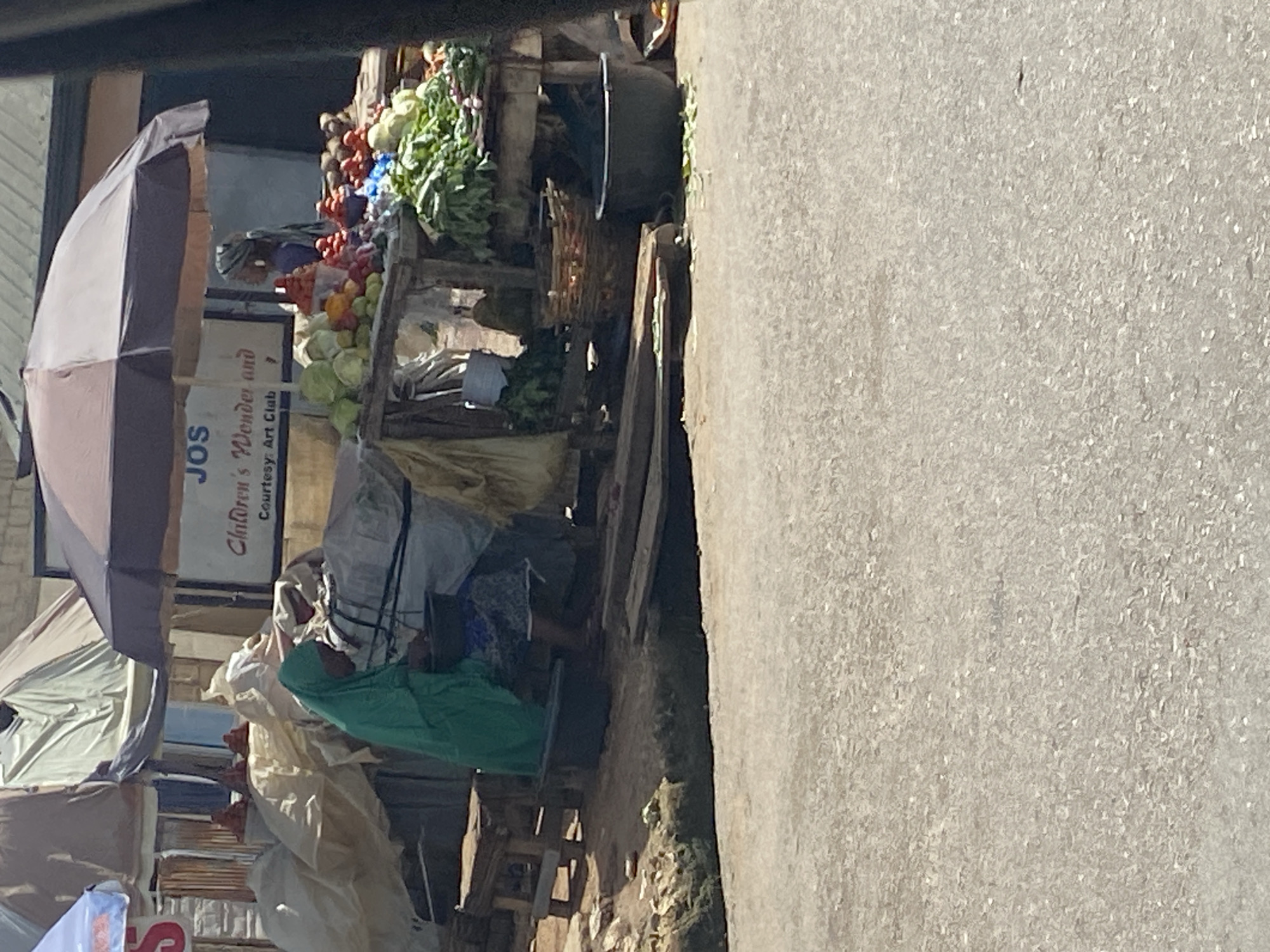 This image depicts an outdoor market scene in Jos, Nigeria. In the foreground, a paved road is visible. Behind the road, a market stall is set up under a large umbrella, with various fresh produce displayed on wooden tables. Vegetables such as cabbage, tomatoes, and leafy greens are prominent. A woman wearing a green headscarf and blue patterned clothing is seated behind a stall, seemingly engaged in market activities. Several sacks and baskets are present, along with some wooden crates. The background shows a building with a sign that reads "JOS Children's Wonder" and "Courtesy: Art Club." Tents and other market structures are also visible, suggesting a bustling commercial area. The overall environment appears to be a typical urban marketplace in Nigeria.