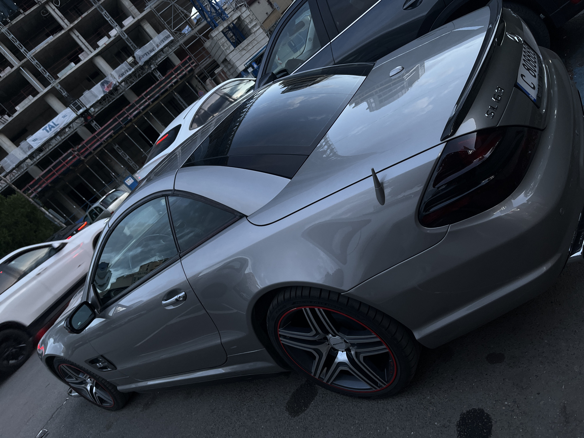 The image captures a silver Mercedes-Benz SL63 AMG parked on an asphalt surface. The car's rear end is prominently featured, showing details like its carbon fiber spoiler, tinted taillights, and a European license plate with "C 8088BB" visible. The distinctive AMG wheels with red accents are also noticeable.
In the background, a building under construction with exposed scaffolding and banners (possibly related to "TAL" and "APTEK") is visible, providing an urban context. Other vehicles, including a white car, are parked nearby.
Based on the license plate, the location is likely Sofia, Bulgaria. The scene appears to be a typical urban street setting with ongoing construction activity in the vicinity.