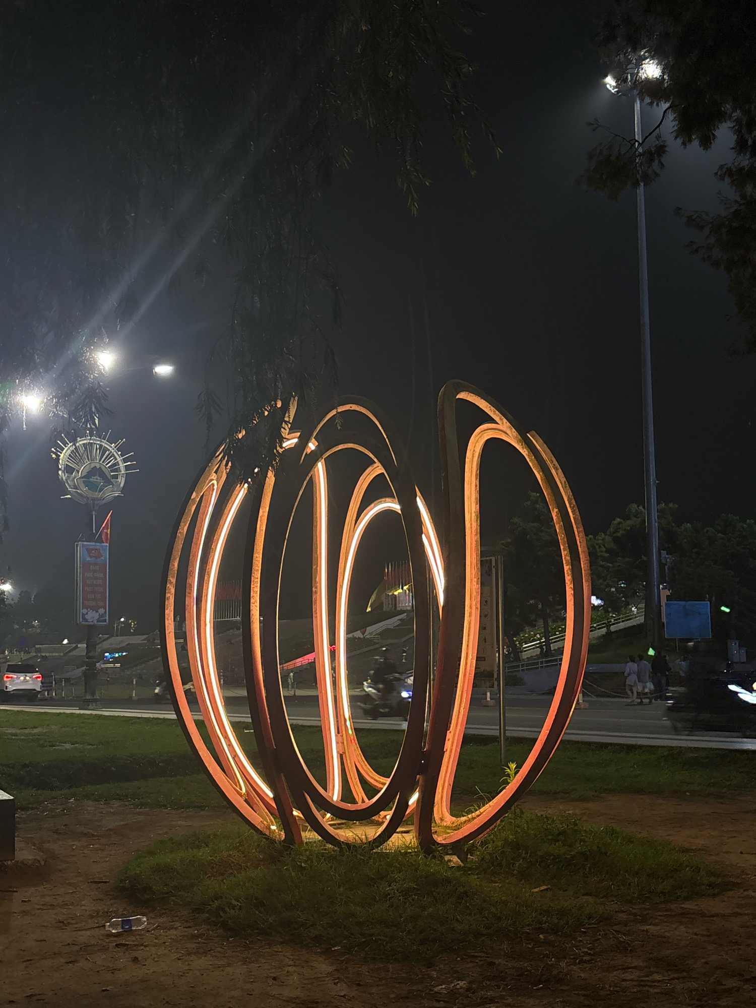 An abstract, circular sculpture illuminated with warm, orange light stands in a grassy area at night. The sculpture is made of several curved metal rings, some of which are attached to each other, creating an open, layered effect. Behind the sculpture, a street is visible with cars and motorcycles passing by, as well as a lamppost and trees. In the background, there are buildings and a sign with a circular emblem. The ground in front of the sculpture is covered with dirt and grass, and a plastic bottle lies on the ground. The overall scene suggests an outdoor urban setting at night.
